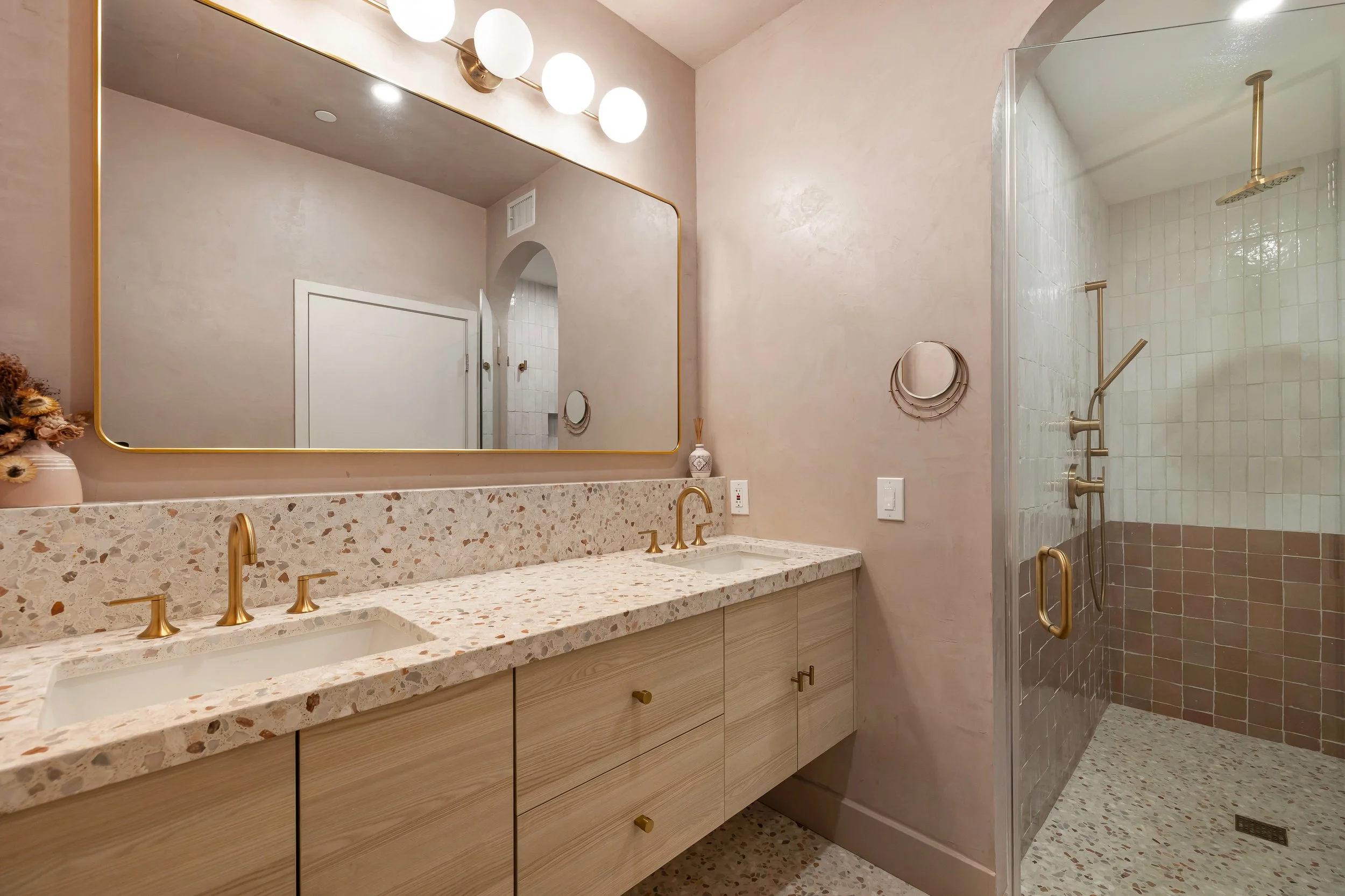 A modern bathroom with a large mirror, a terrazzo countertop with a double sink and gold fixtures, beige cabinetry, and a walk-in shower with a glass door and gold fixtures.
