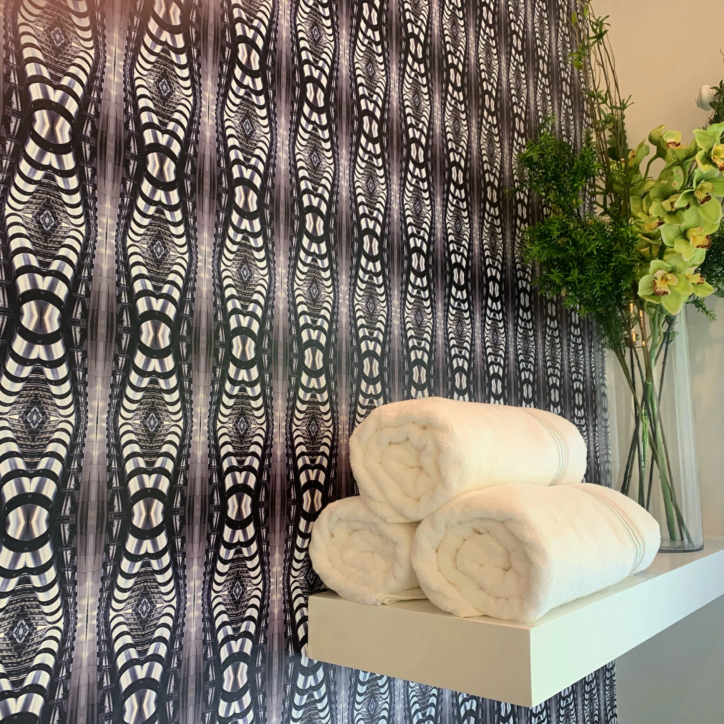 Three rolled white towels placed on a white shelf next to a large vase with green and yellow flowers and a tall green plant, against a patterned black and white curtain background.