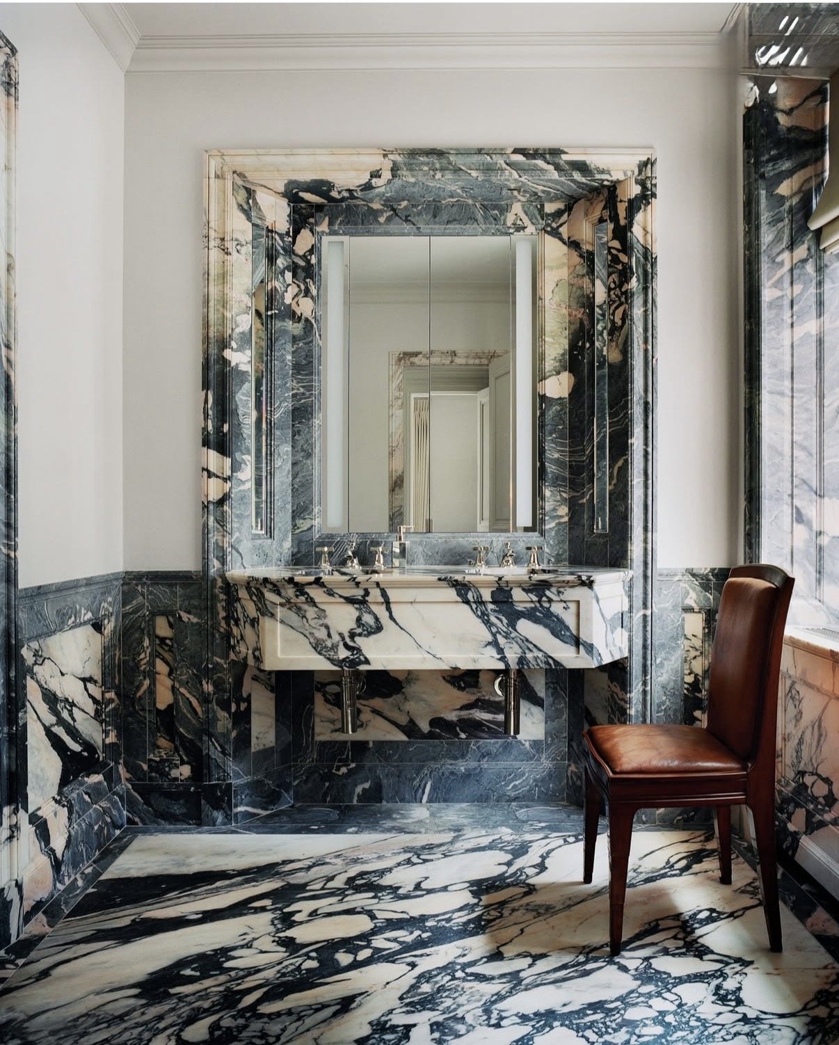 A luxurious bathroom with black and white marble walls, floor, and a marble vanity with a large mirror, and a brown leather chair by a window.
