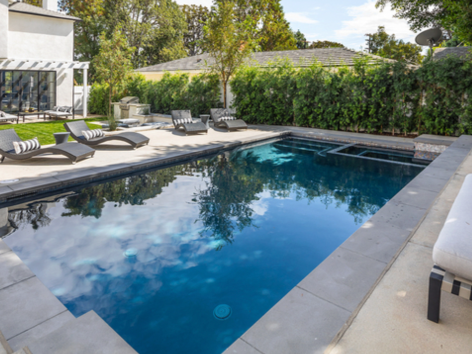 Backyard swimming pool with lounge chairs, surrounded by greenery and a house with large windows.