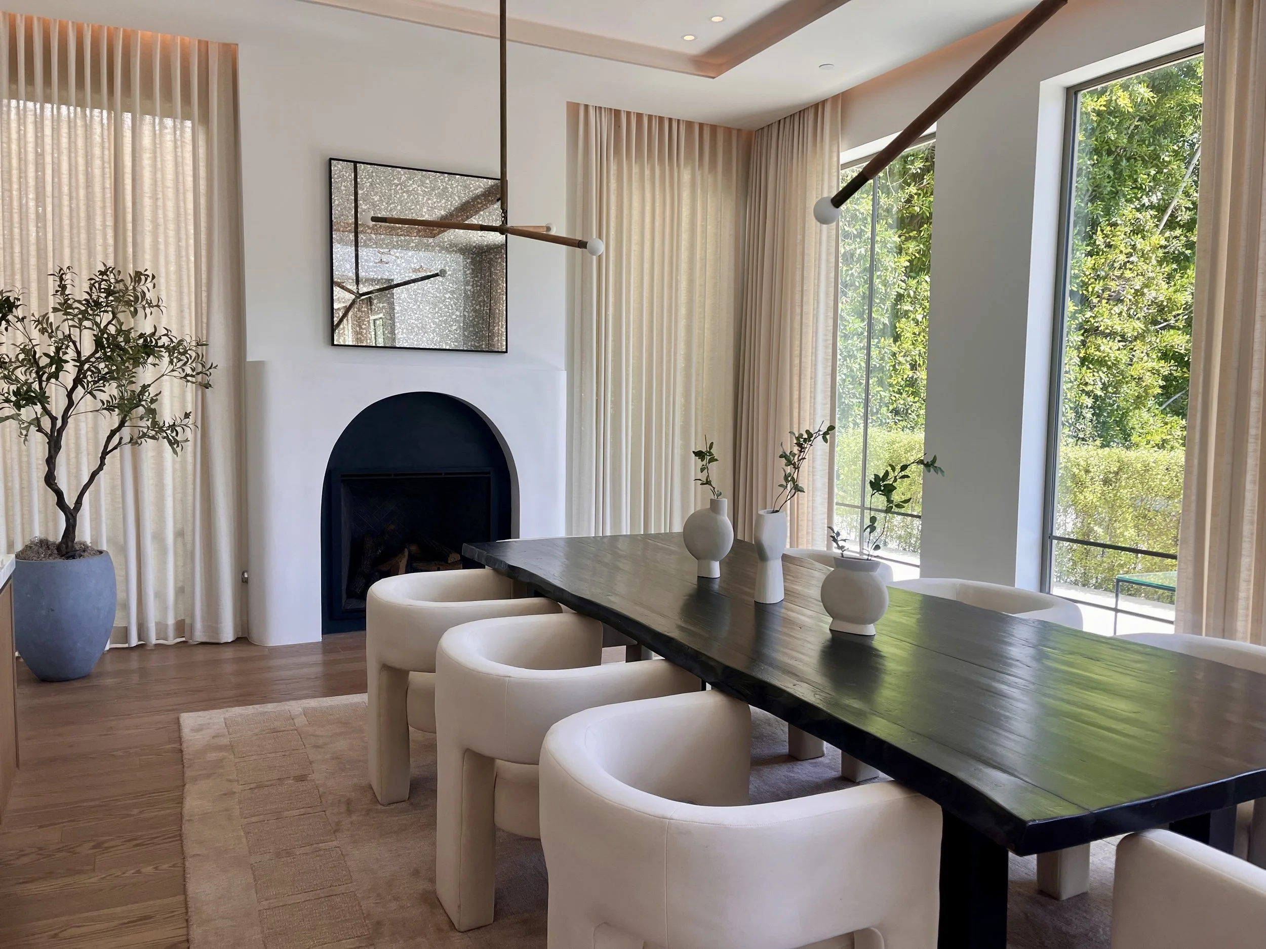 Modern dining room with a dark wooden table, six cream-colored upholstered chairs, three white vases with green branches, large floor-to-ceiling windows with beige curtains, and a fireplace with a mirror above it.