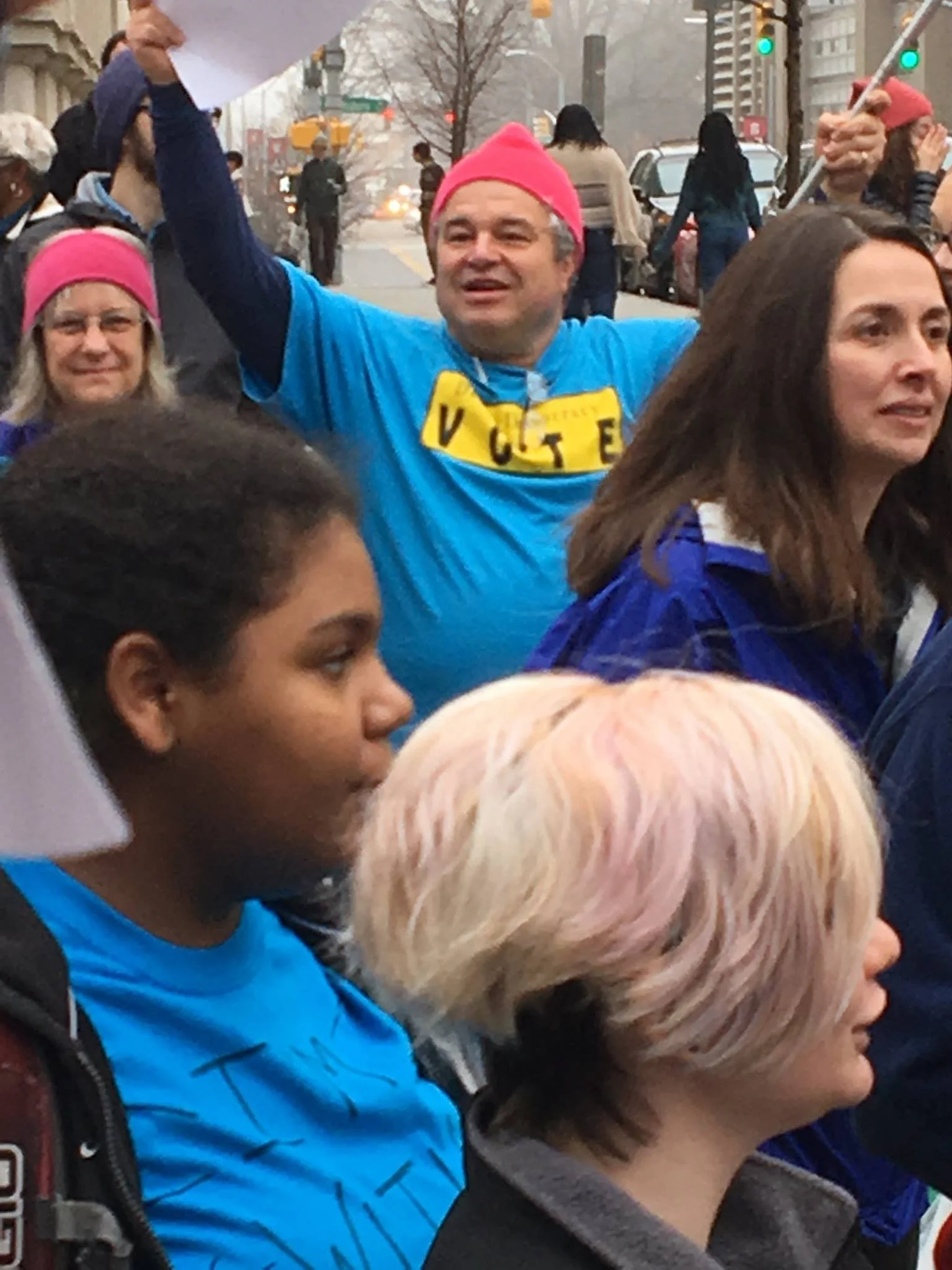 A crowd of people participating in a street protest or rally. One man in the center wearing a pink hat and blue shirt with yellow letters celebrating 'VOTE' is raising his arm, holding a piece of paper. Several women are visible around him, some also wearing pink hats, and the scene takes place in an urban setting with traffic lights and buildings in the background.