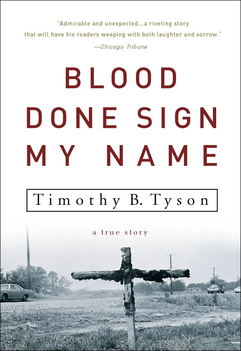 Book cover of 'Blood Done Sign My Name' by Timothy B. Tyson, featuring a black and white photo of a rural landscape with a weathered wooden cross and cars in the background.