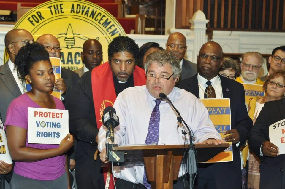 A group of people gathered around a man in a white shirt and purple tie speaking at a podium. Some hold signs with messages like 'Protect Voting Rights' and 'Stop Voter Ejection.' There is a large yellow banner in the background with the words 'For the Advancement' visible.