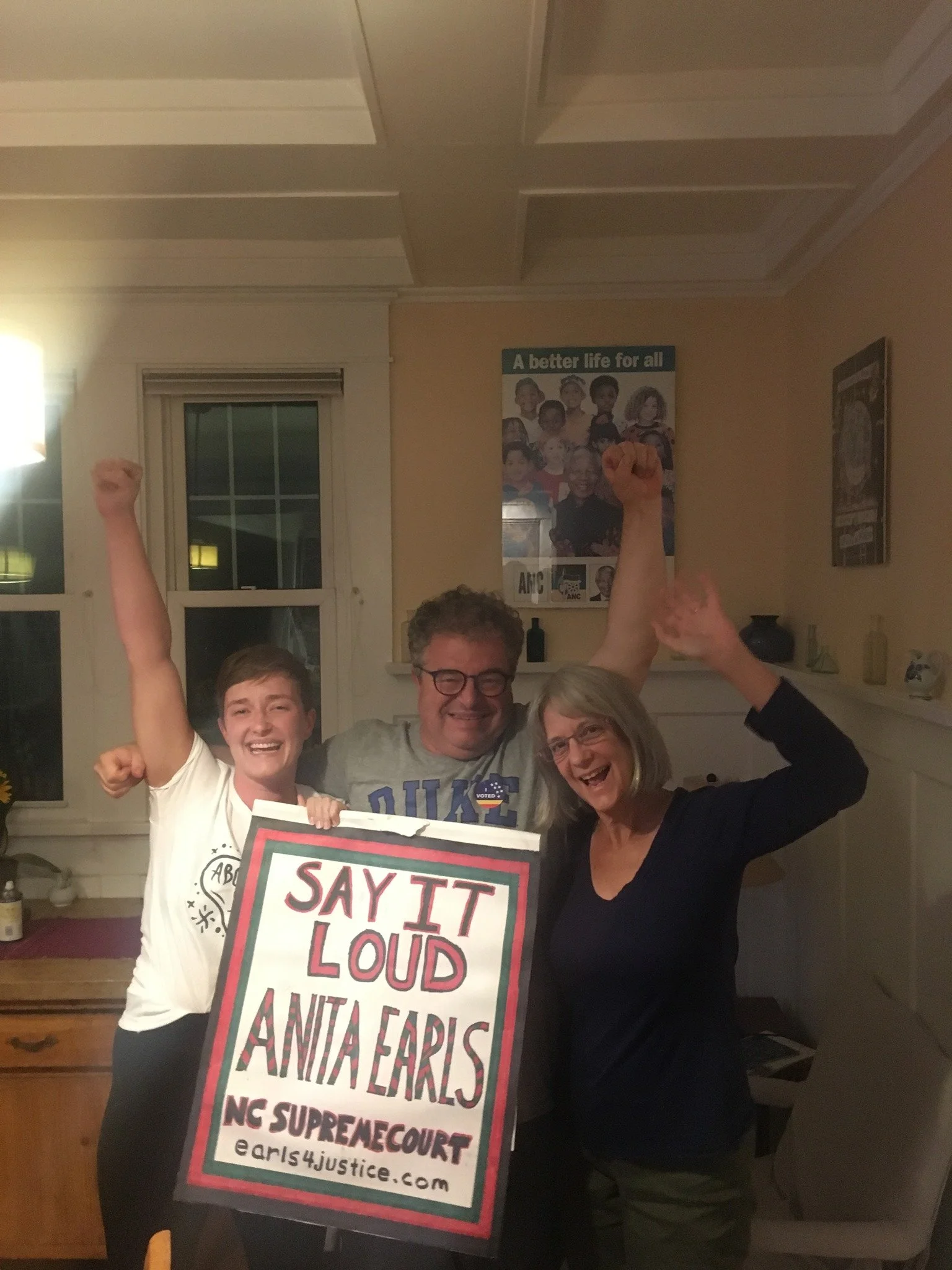 Three people celebrating indoors, holding a sign that says 'Say It Loud Anita Earls NG Supreme Court' with their arms raised and smiling.