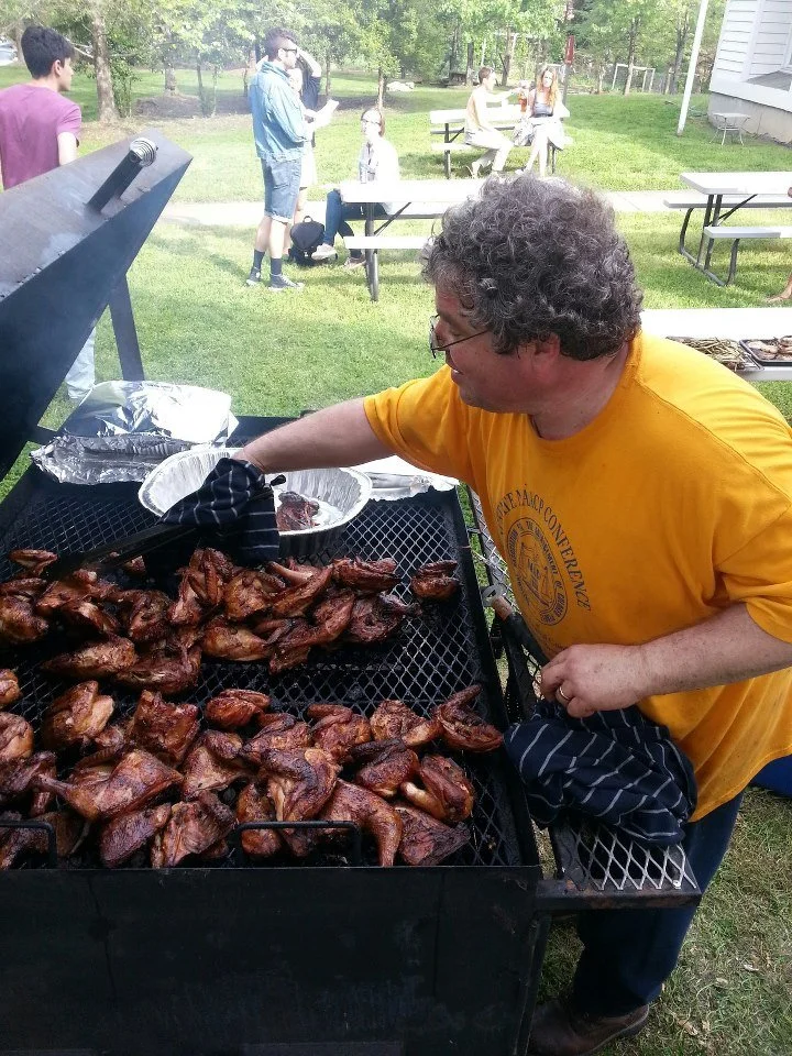 Man in yellow shirt grilling chicken wings at outdoor barbecue with several people in the background