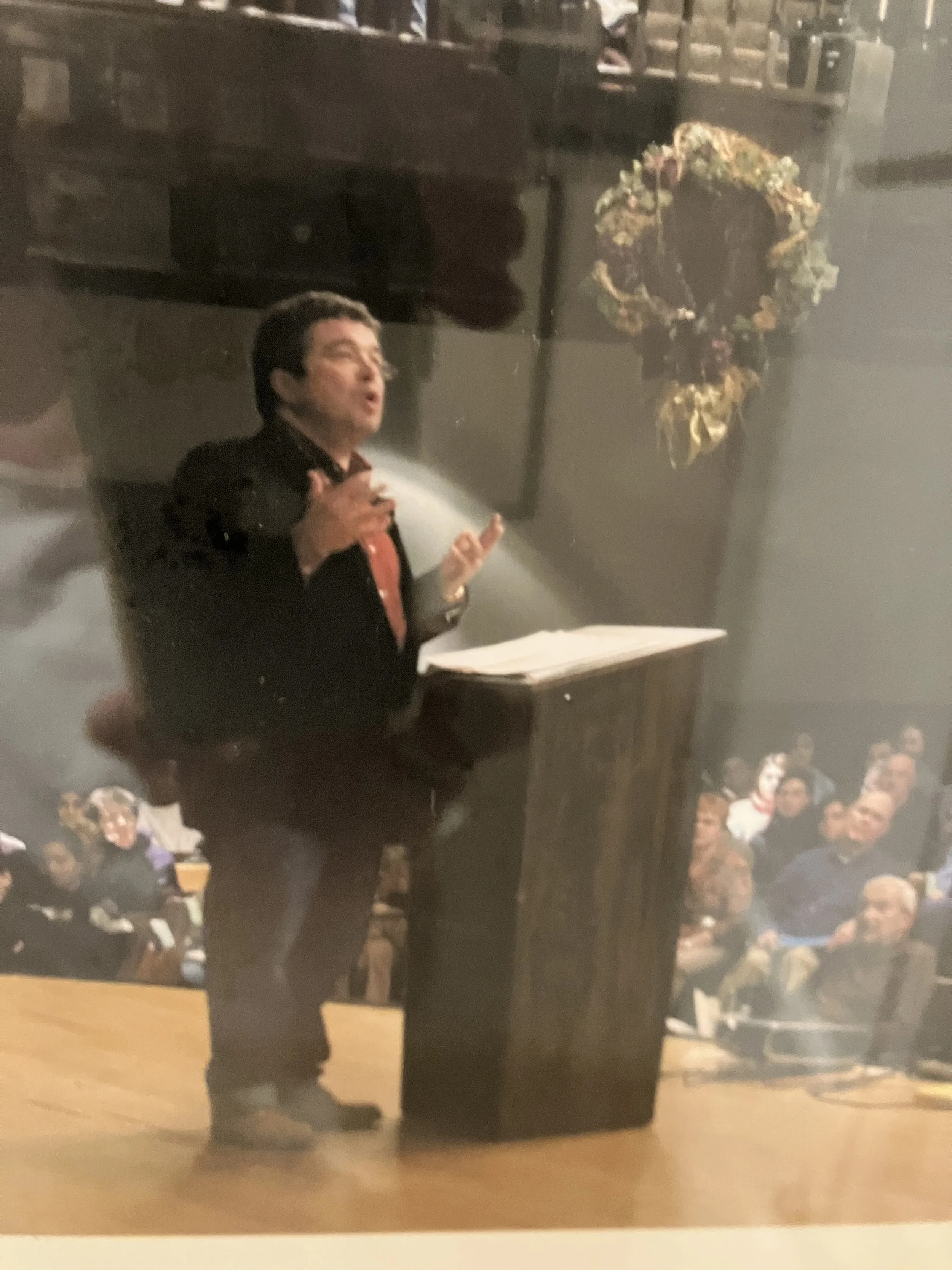 A person standing at a podium speaking to an audience in a large room with a floral wreath hanging on the wall behind them.