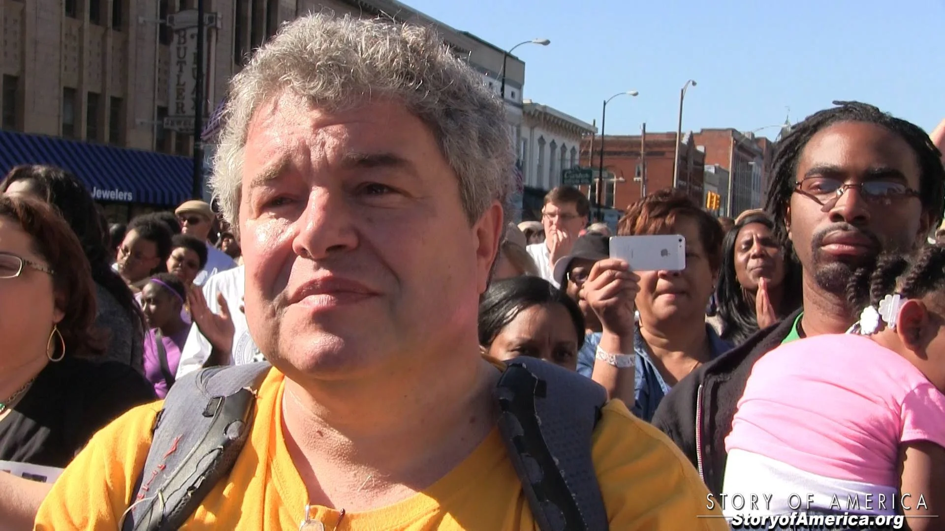 Crowd of diverse people gathered outdoors on a sunny day, some taking photos or videos, with buildings and a blue sky in the background.