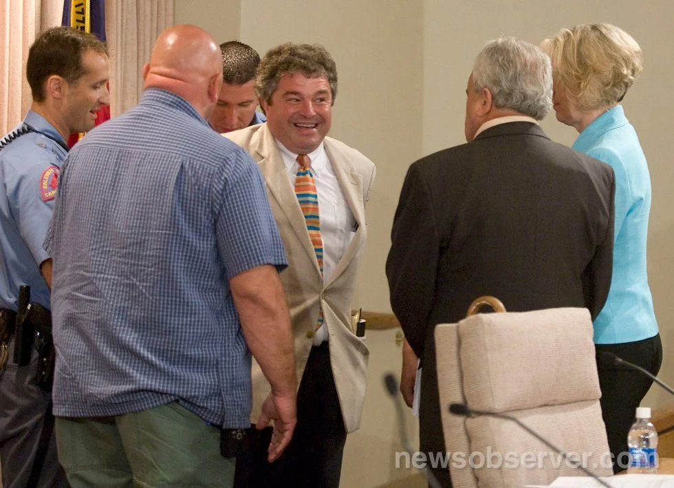 Six people, including two police officers and four civilians, are engaged in conversation and smiling in a room with beige walls. One man in a tan coat and the others in formal or casual attire.