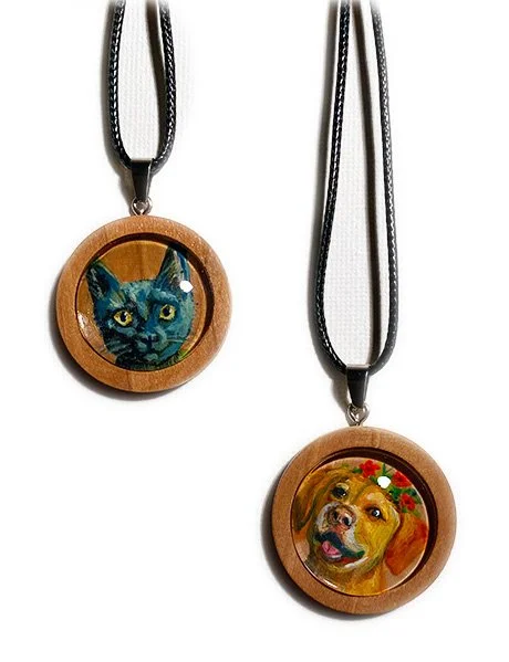 Two wooden pendant necklaces with painted portraits, one of a black cat with yellow eyes and the other of a brown dog with a red and green floral collar, hanging on black cords.