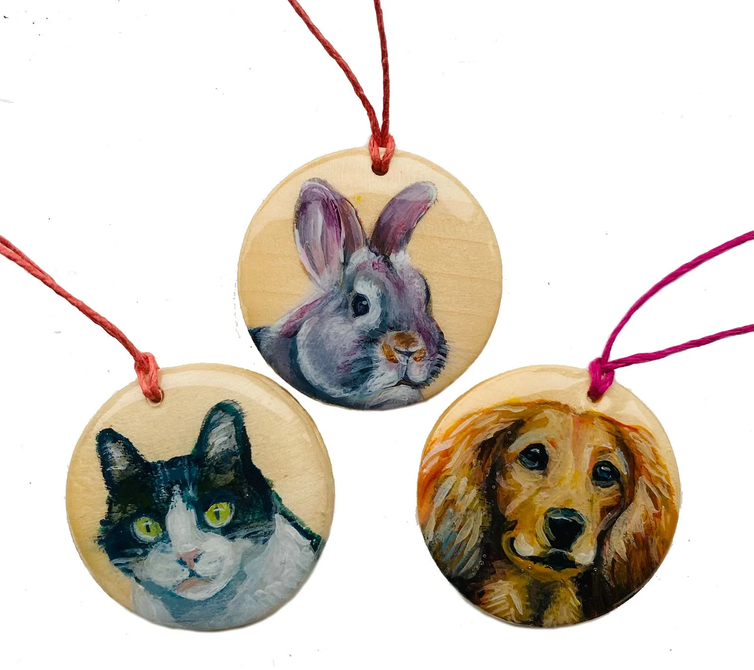 Three round wooden ornaments with painted animal portraits: a rabbit in the center, a cat on the left, and a dog on the right, each with a string for hanging.