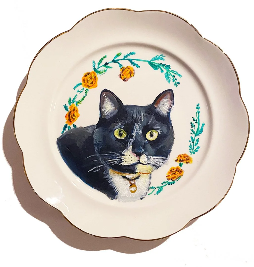 Decorative plate featuring a hand-painted portrait of a black and white cat with yellow eyes, surrounded by a floral wreath of orange flowers and green greenery.