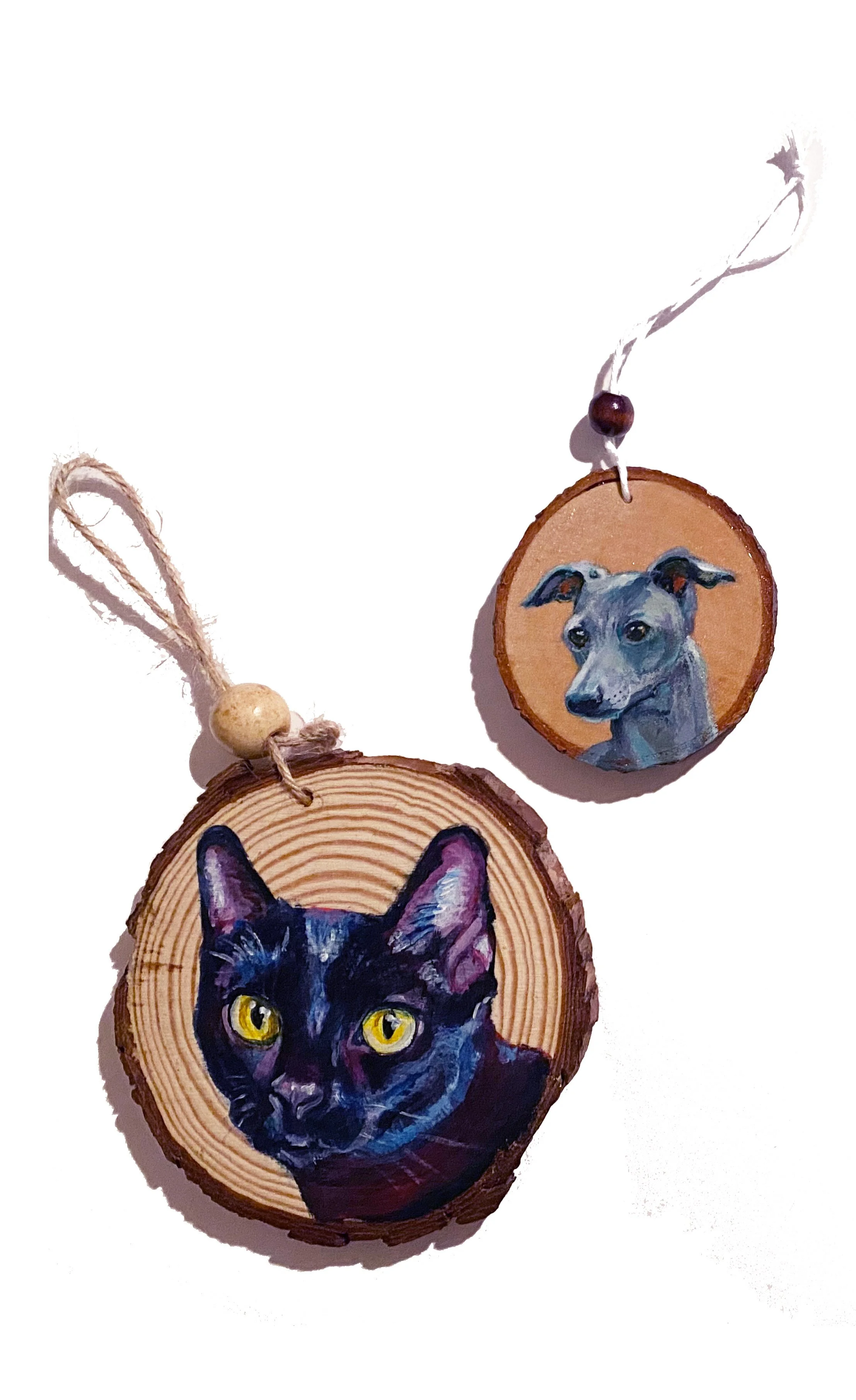 Painted animal portrait ornaments on wood slices, one with a black cat with yellow eyes and the other with a gray dog, both with hanging strings.