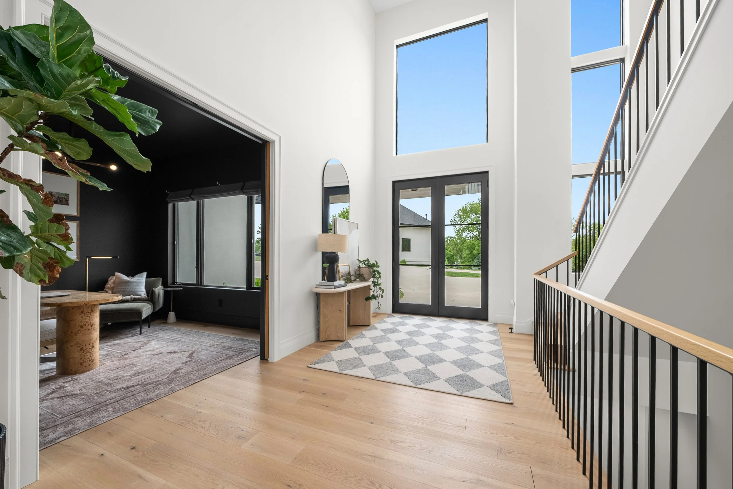 Modern entryway with light wooden flooring, large windows, black front door, and a staircase with black railing. Small wooden console table with a lamp and plant. Rug with a geometric pattern near the door. Adjoining room with dark walls and furniture.