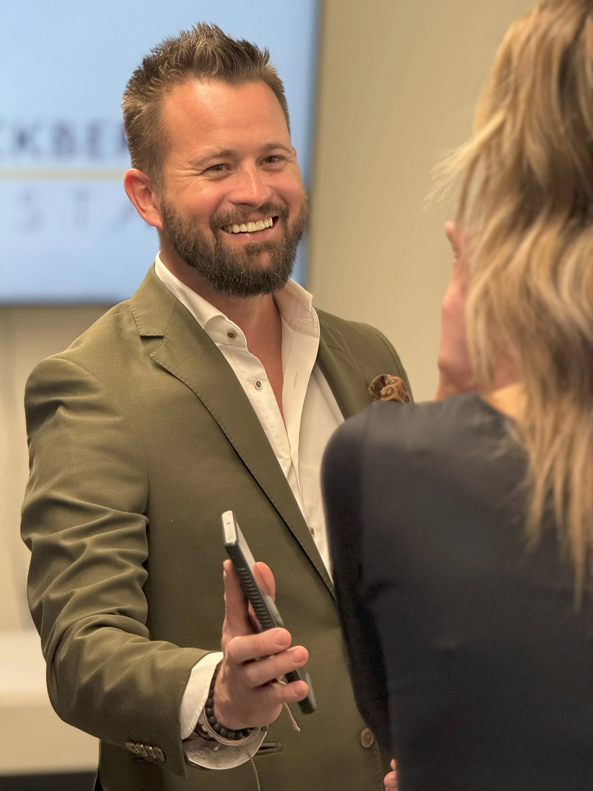 A man with a beard and short hair in a green blazer smiling at a woman during a conversation in an indoor setting, with a screen in the background.