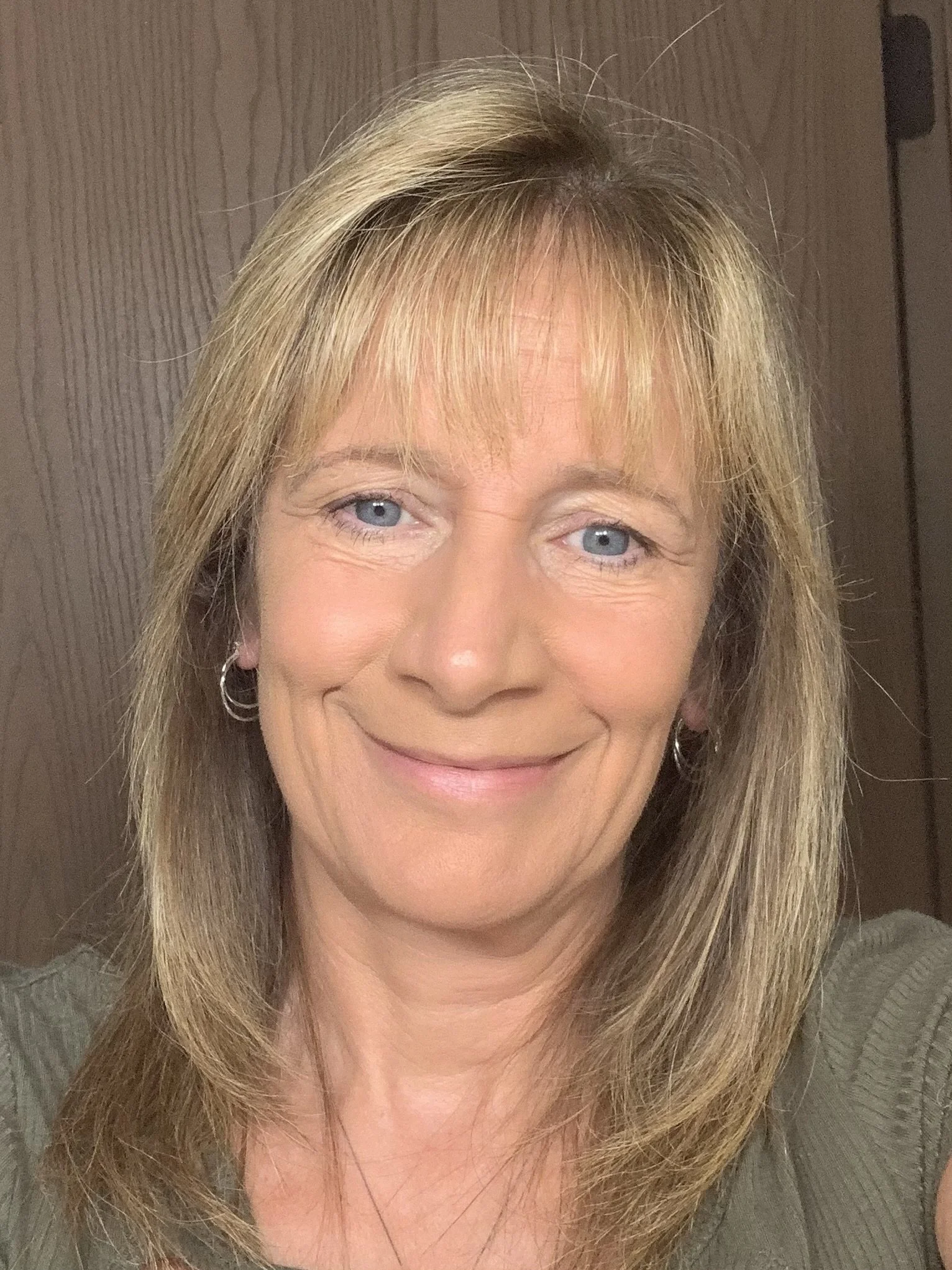 Close-up of a woman with blonde hair and blue eyes, smiling, in front of a wood-paneled wall.