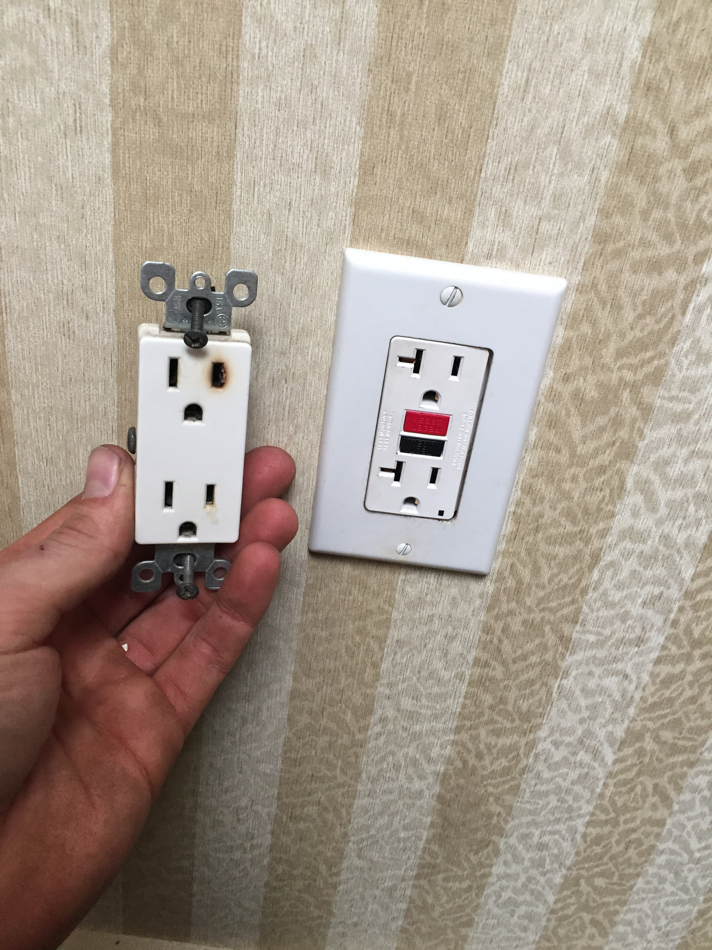 Hand holding a removed electrical outlet cover next to a wall socket with a circuit breaker switch attached.