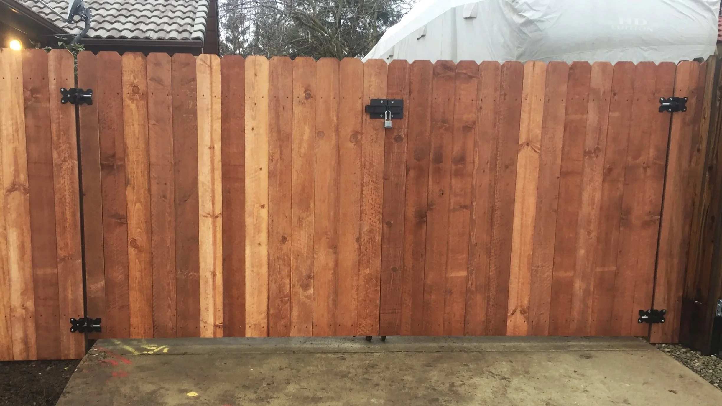 A wooden gate with a latch lock, outside on a patio, with a house and trees visible in the background.