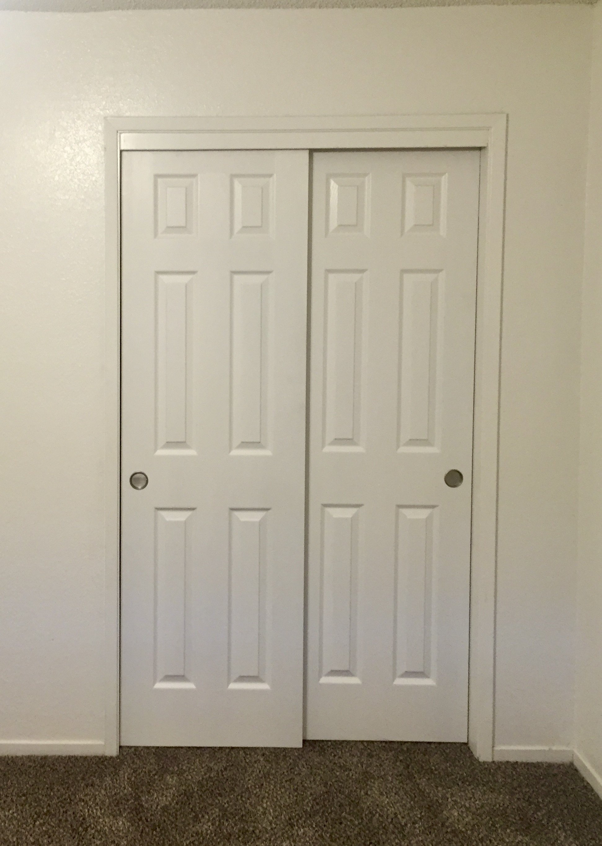 White double sliding closet doors on a wall, with a strip of trim around the frame, set against a white wall with a textured surface, and a brown carpeted floor.