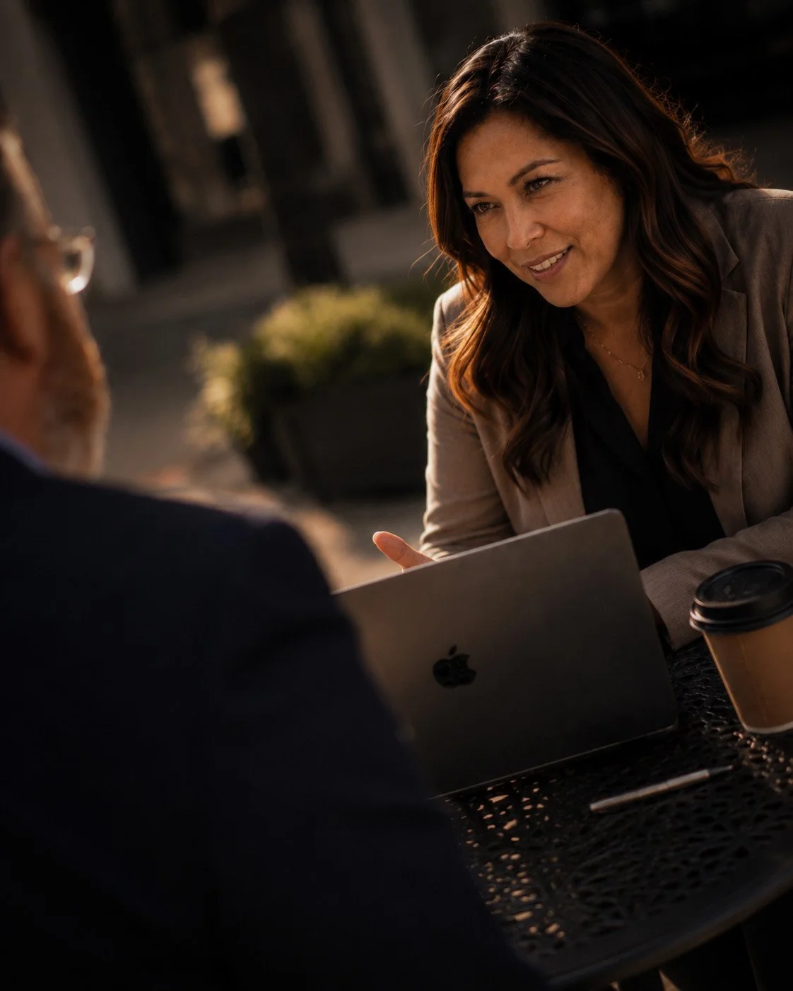 A woman with brown hair smiling and engaging in conversation with a man, sitting at an outdoor table with a coffee cup and a laptop.