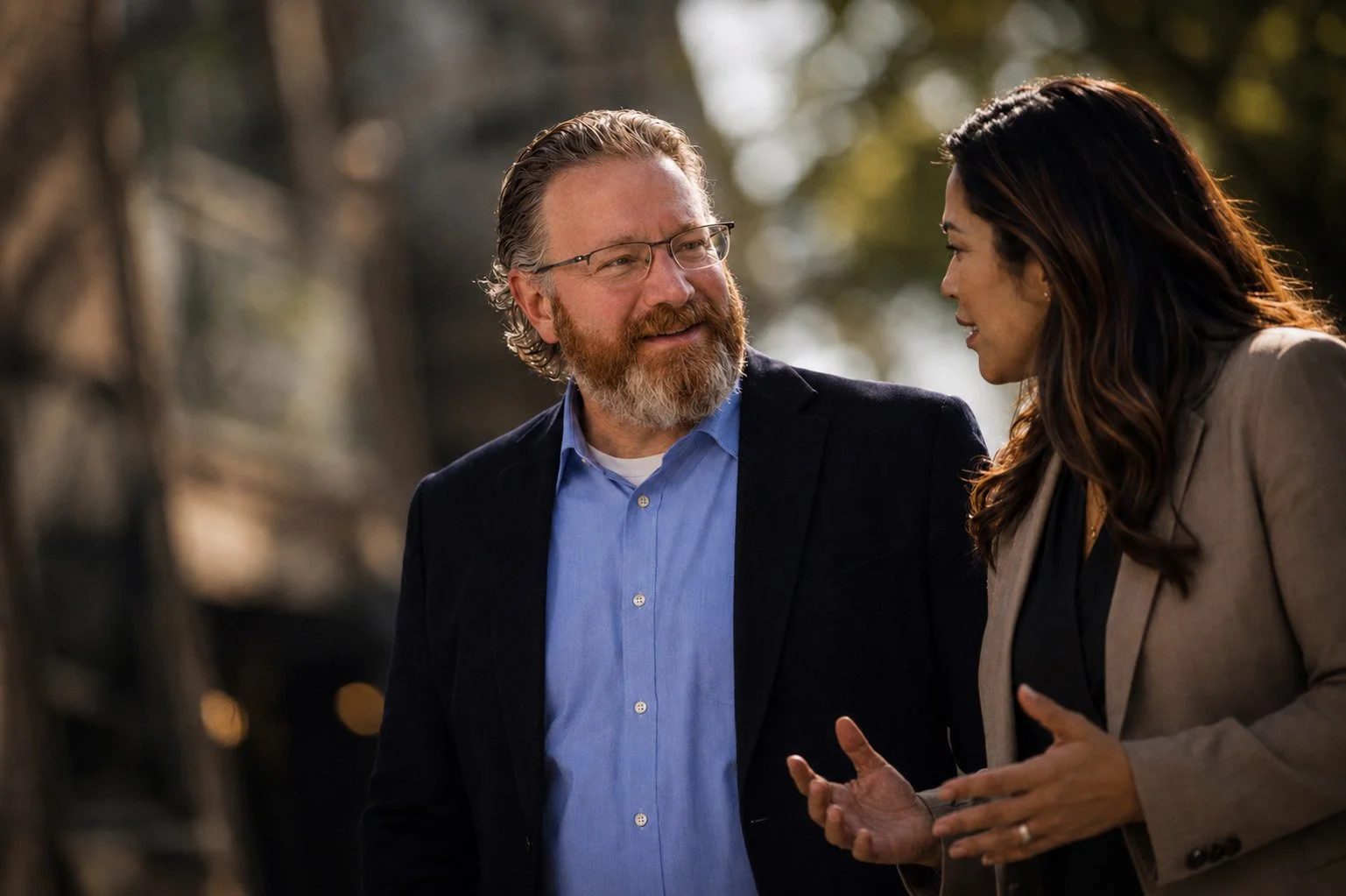 A man with glasses and a beard in a dark blazer talking to a woman with long dark hair in a beige blazer outdoors during daylight.
