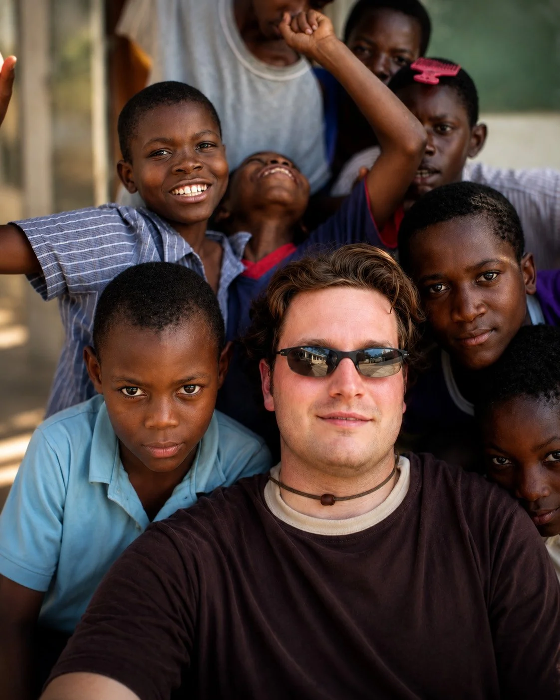 A younger Timothy San Fillippo surrounded by smiling Haitian children outside a weathered school building during his 2004 service in Grand Goâve, Haiti.