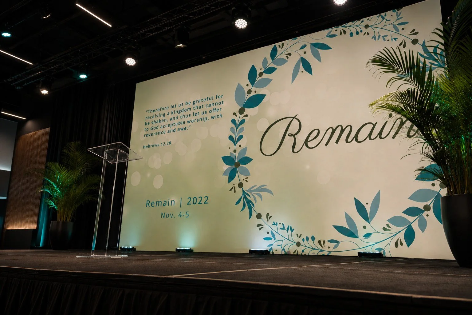 Remain 2022 women’s conference stage with a floral Scripture backdrop, acrylic podium, green plants and teal lighting.