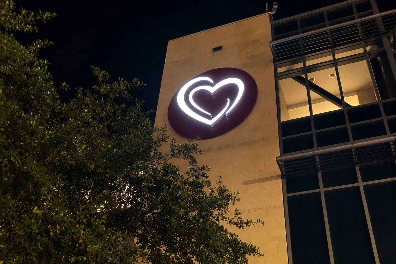 Nighttime exterior view of a building with a large illuminated heart logo on the wall, partially obscured by tree branches.