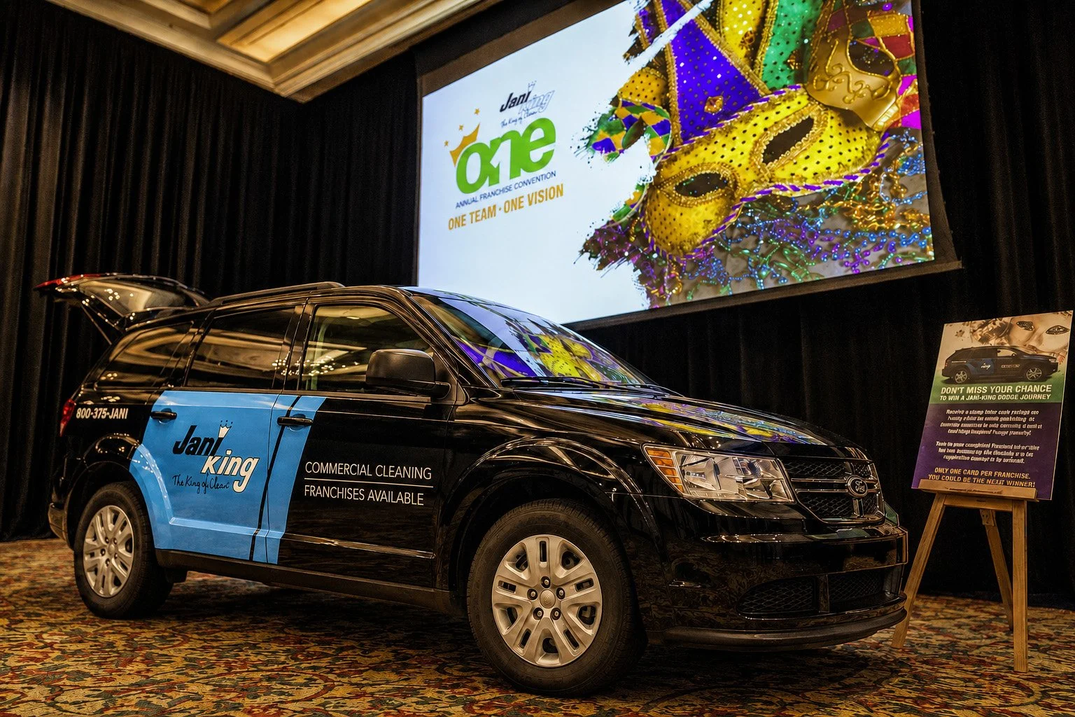 Black Dodge Journey with Jani-King Gulf Coast graphics displayed beneath a Mardi Gras-themed ONE convention screen.