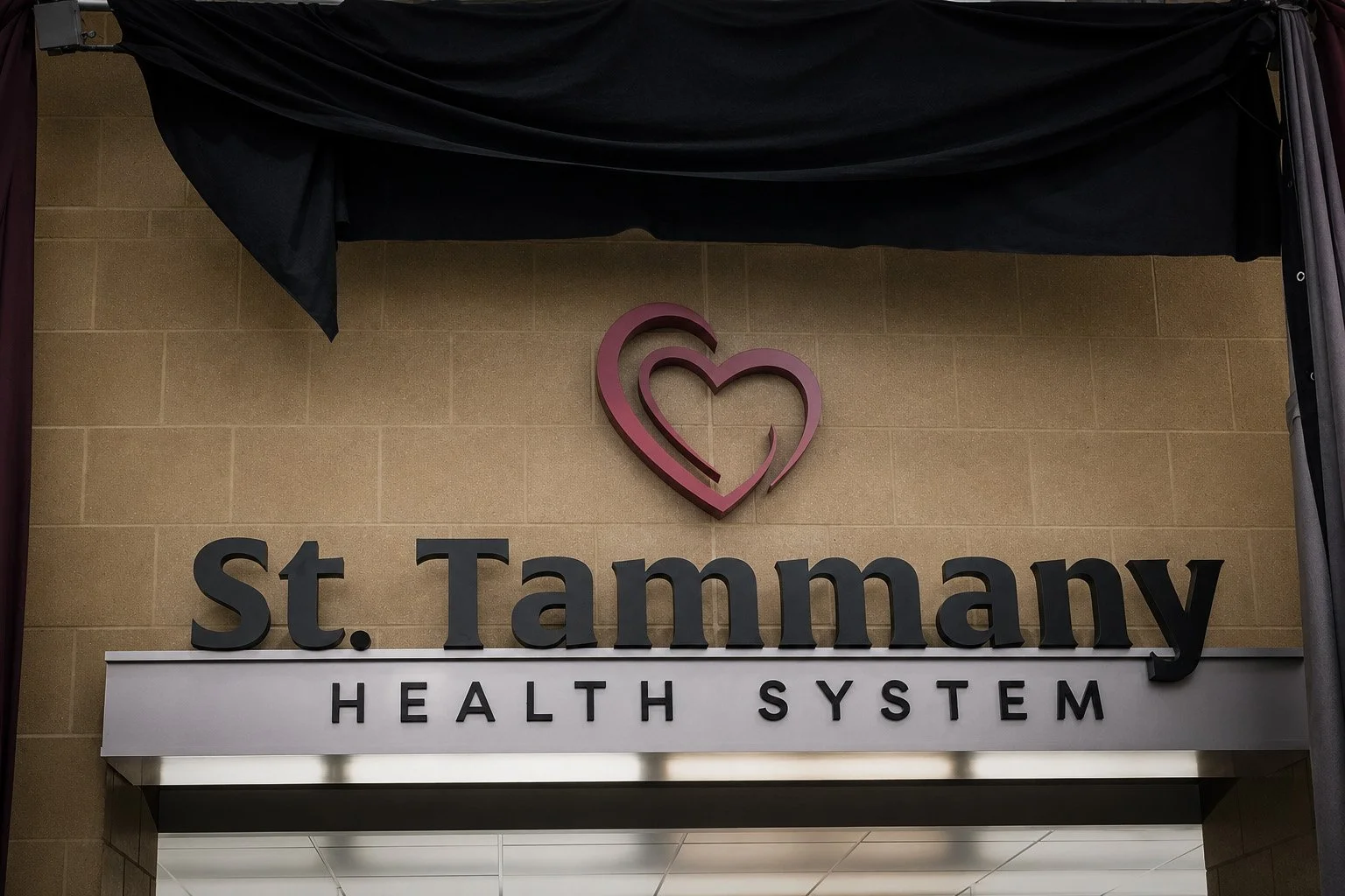 St. Tammany Health System name and red interlocking hearts displayed on a tan wall beneath black reveal drapery.
