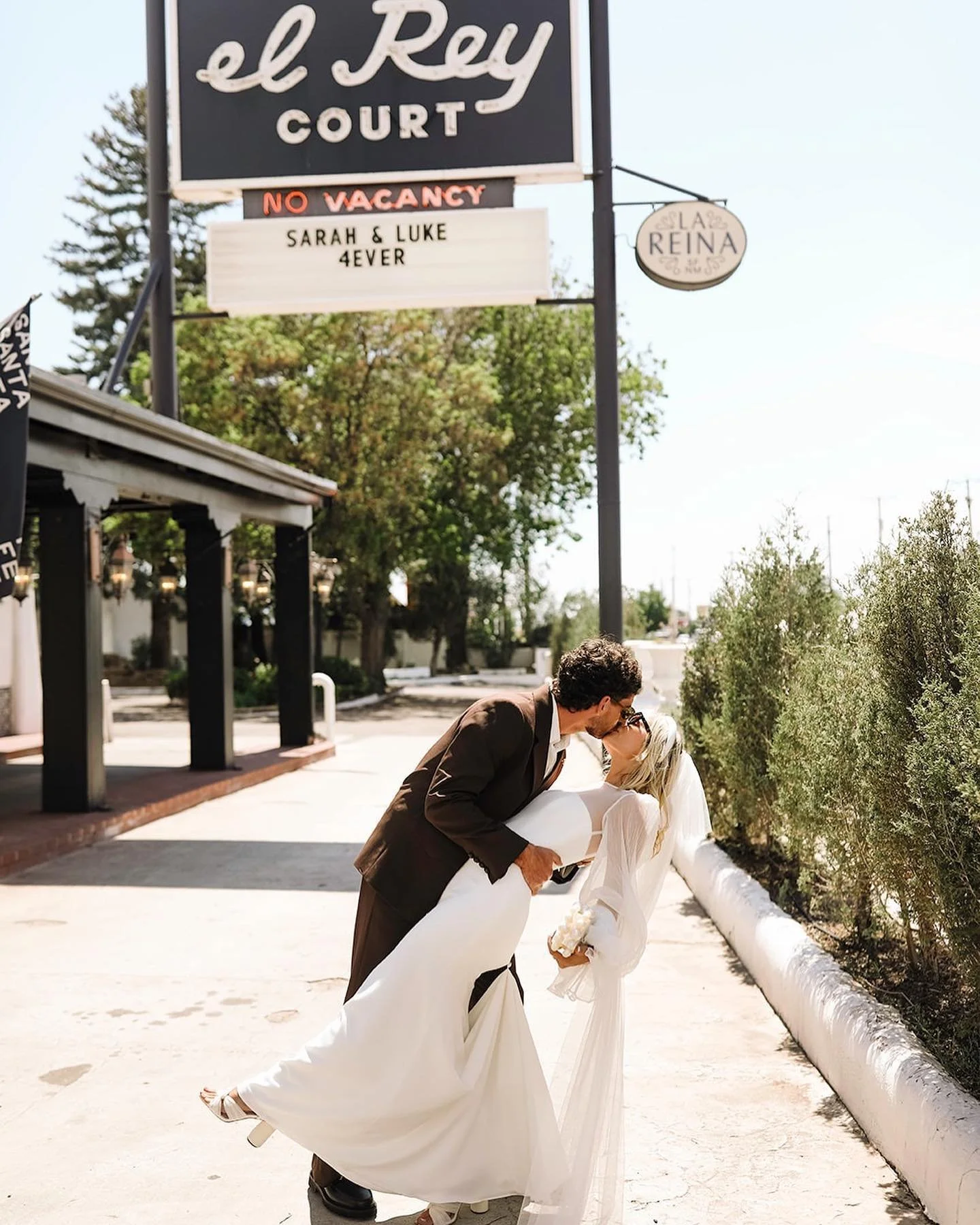 A couple dressed in wedding attire sharing a romantic kiss outdoors, with the groom dipping the bride as they lean over a sidewalk lined with bushes, under a sign for el Rey Court.
