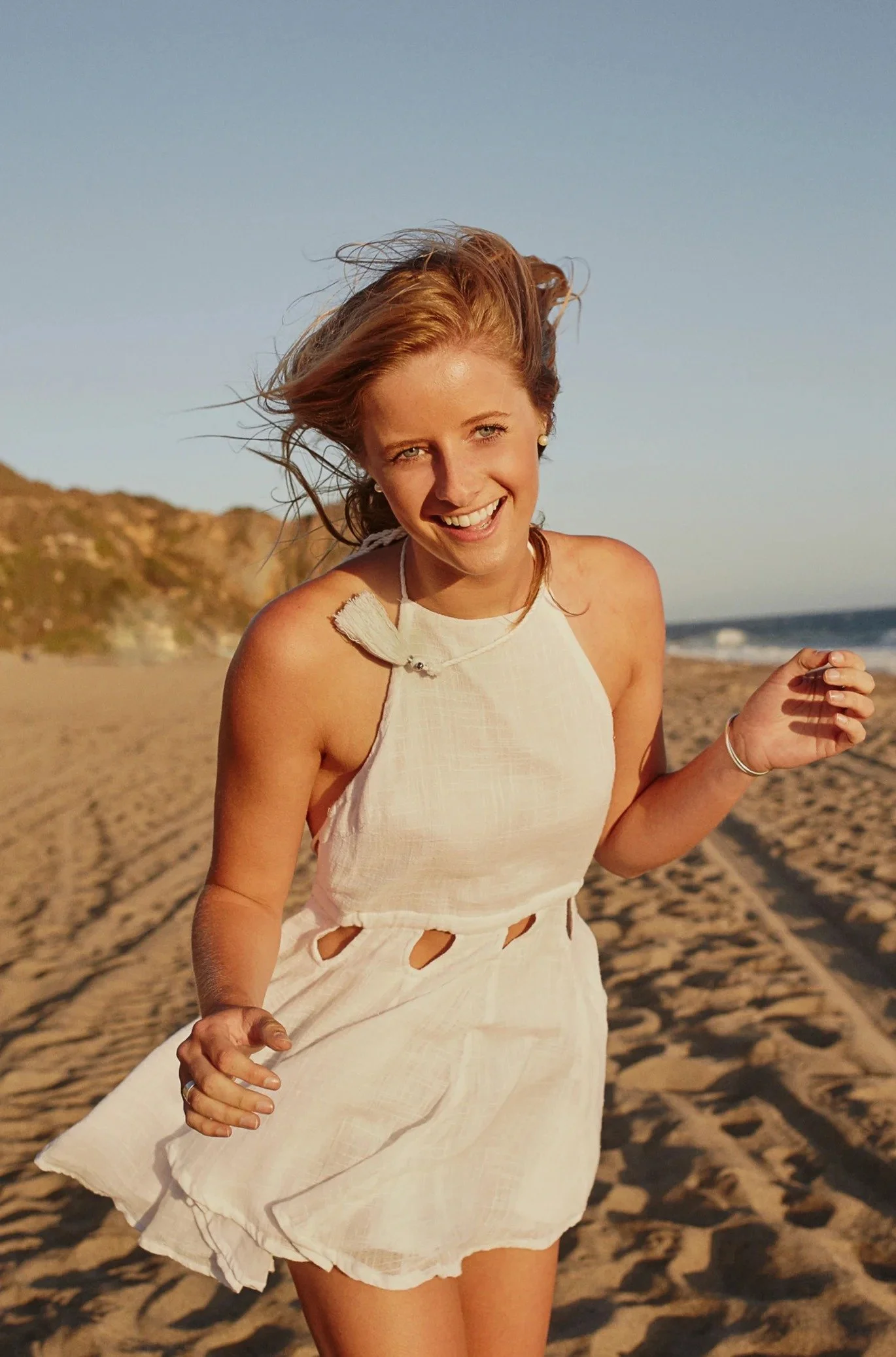 A woman smiling at the beach, wearing a white sleeveless dress with cutouts around the waist, and her hair blowing in the wind.