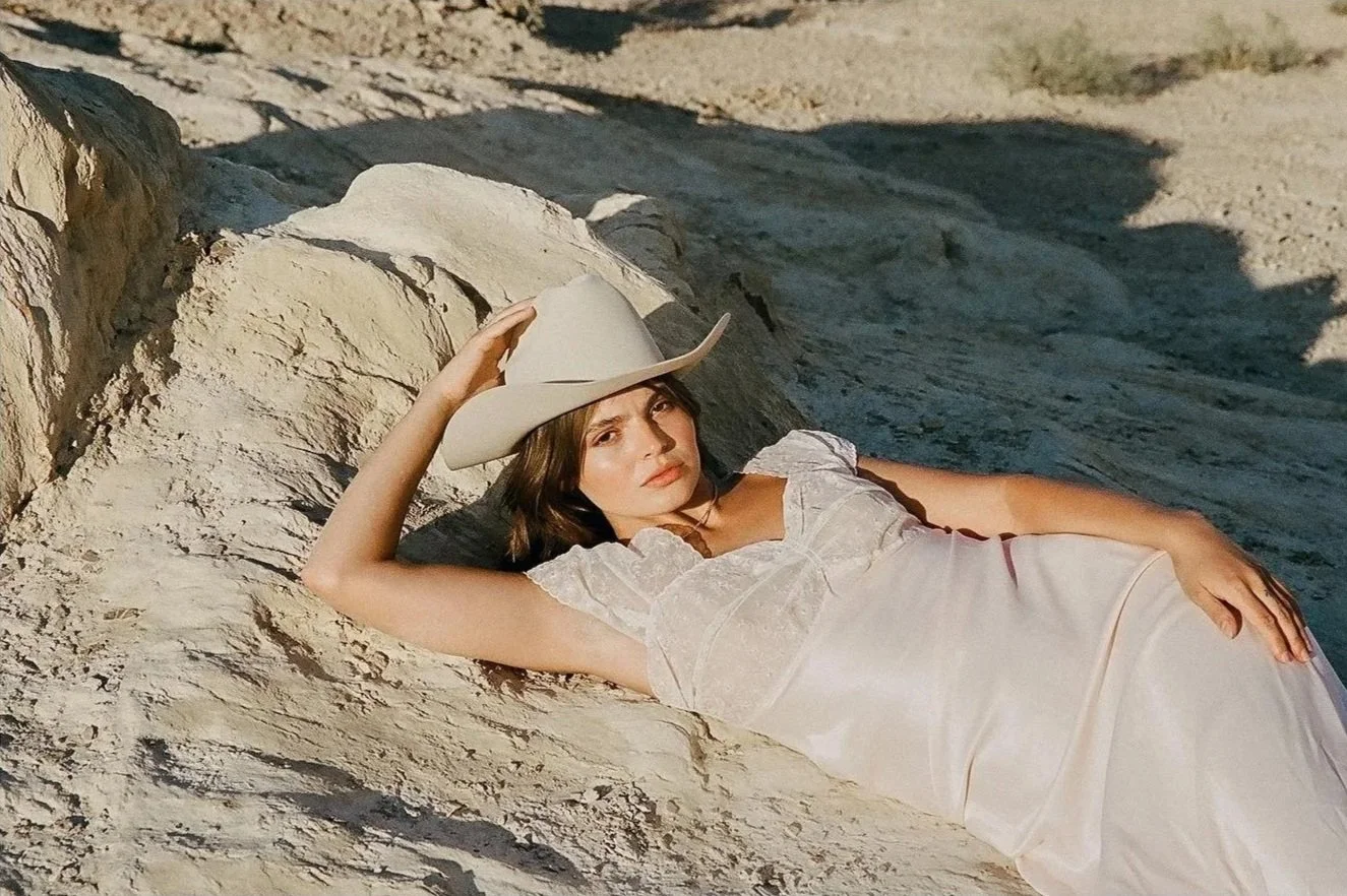 A woman lying on a large rock formation, wearing a white dress and a cowboy hat, in a desert or rocky landscape.