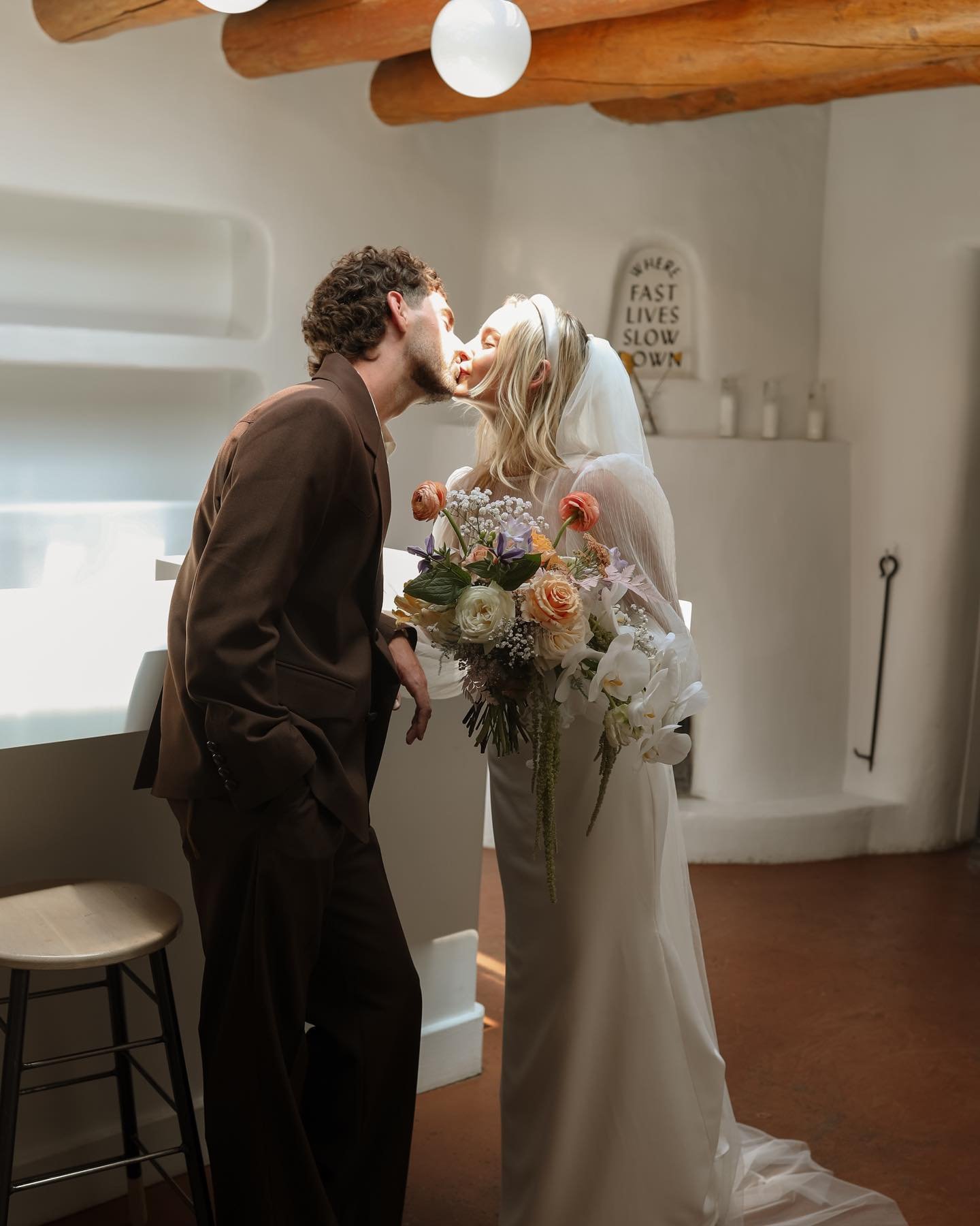 A man and woman, dressed in wedding attire, sharing a kiss indoors. The woman is holding a large bouquet of flowers. The room has a lived-in, cozy feel with wooden beams on the ceiling and decorative signs on the wall.