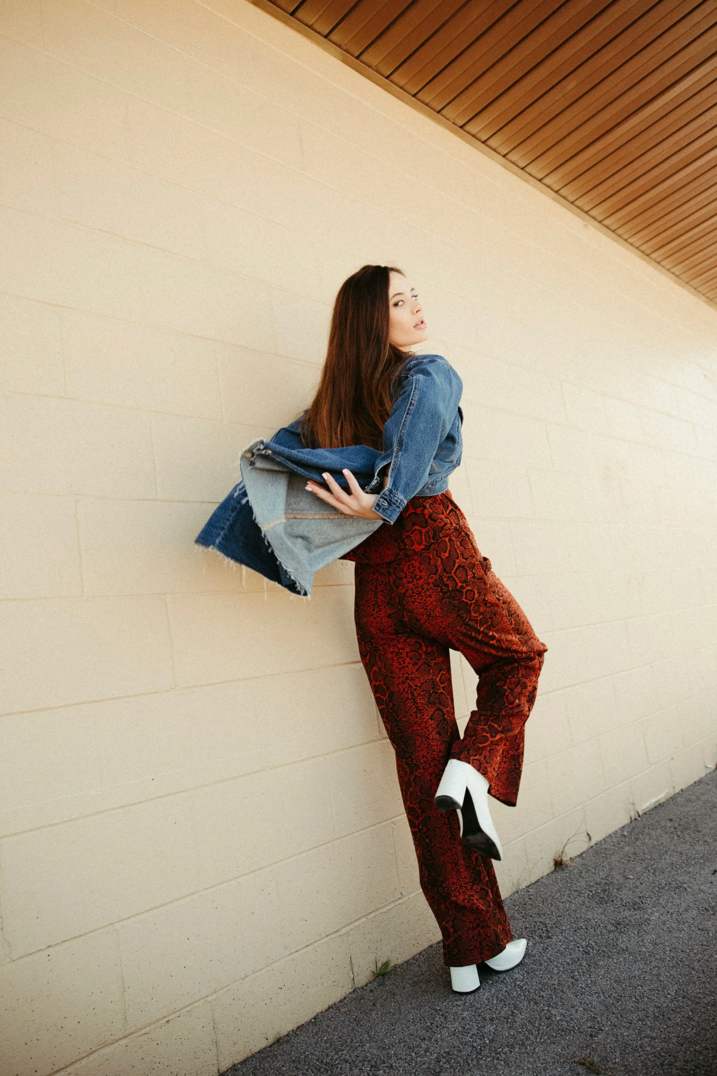 A woman with long brown hair is leaning against a beige wall, wearing a denim jacket draped over her shoulder, a patterned red and black pants, and white platform boots. She looks to her right with a neutral expression.
