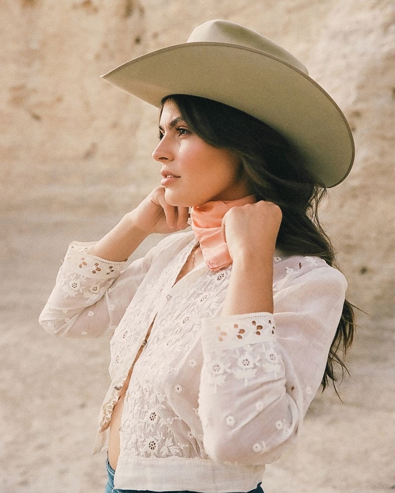 A woman wears a wide-brimmed hat, a white embroidered blouse, and a pink scarf around her neck, posing against a neutral background.