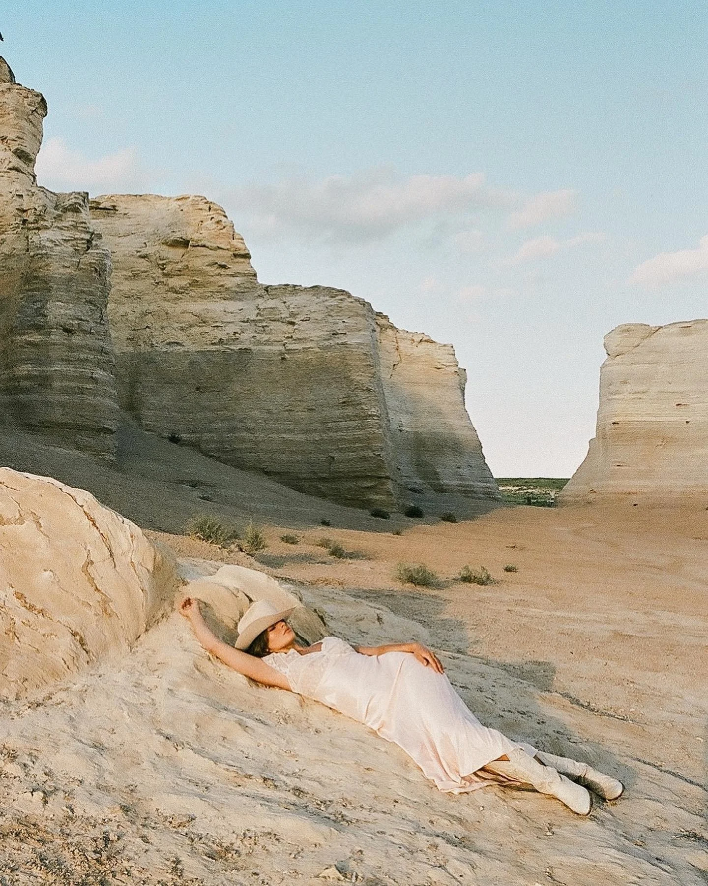 A woman lying on a large rock formation in a desert landscape, wearing a white dress and a wide-brimmed hat, with rocky cliffs and blue sky in the background.
