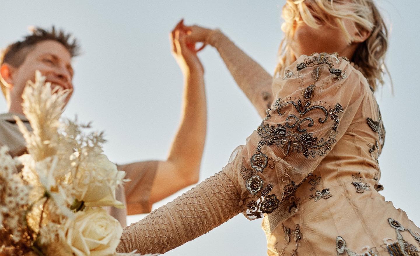 A woman and a man dancing outdoors during sunset, with the woman wearing an embroidered dress and the man smiling near flowers.