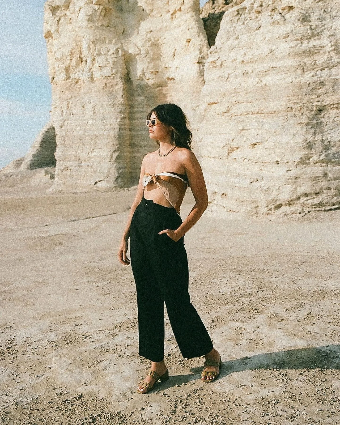 A woman with dark hair, wearing sunglasses, a strapless top, black wide pants, and heels, standing on a sandy beach at the base of tall rocky cliffs.