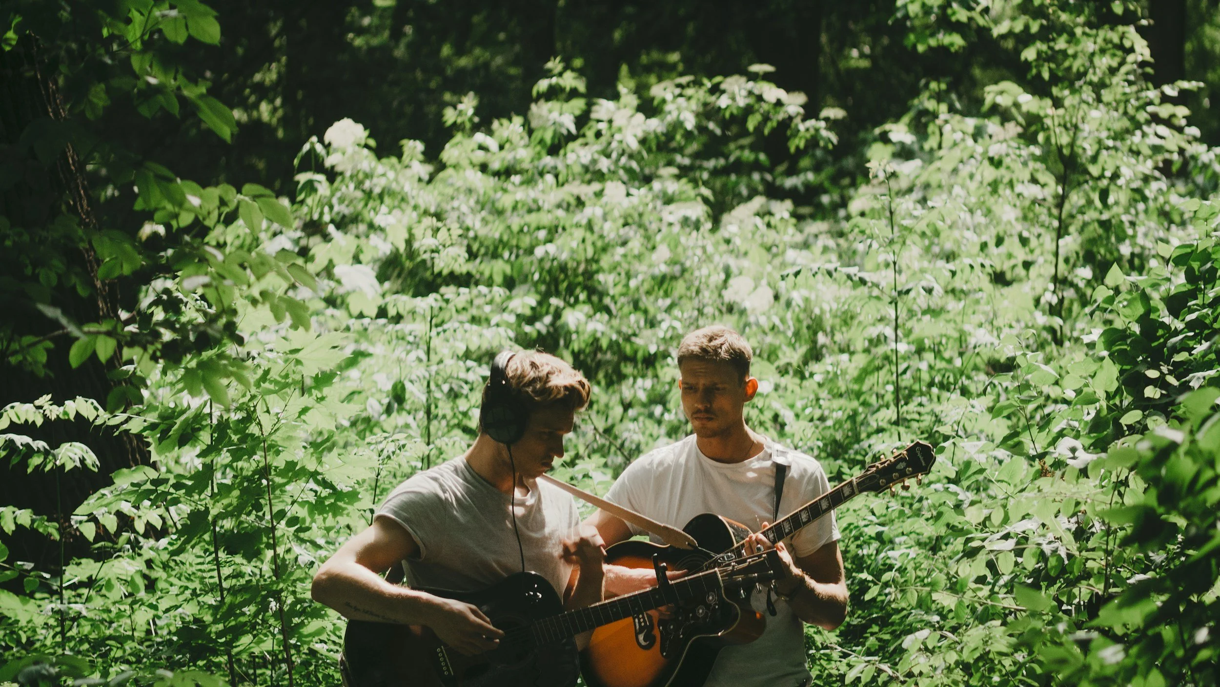 Grounded Media - Two young Musicians playing guitars in a lush green forest, one wearing headphones and the other focused on his guitar.