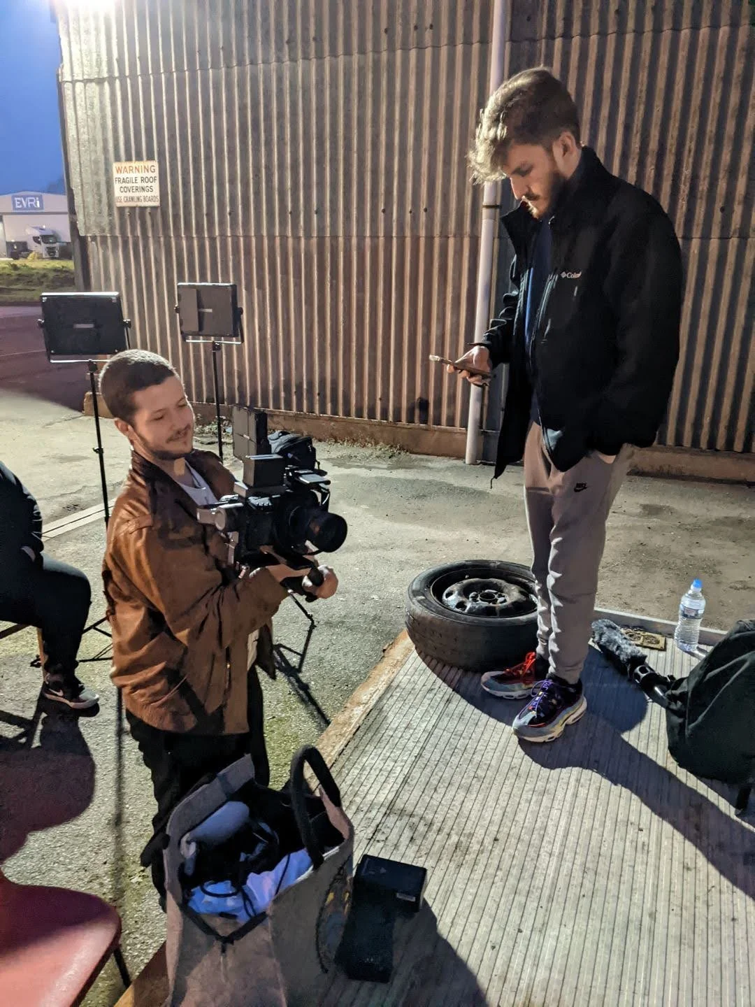Stan Thornton, and a crew of filmmakers, Stan Thornton holding a camera, filming outside at night. One is standing on a wooden platform and the other is looking at his phone. There are lights, a water bottle, and a backpack in the scene.