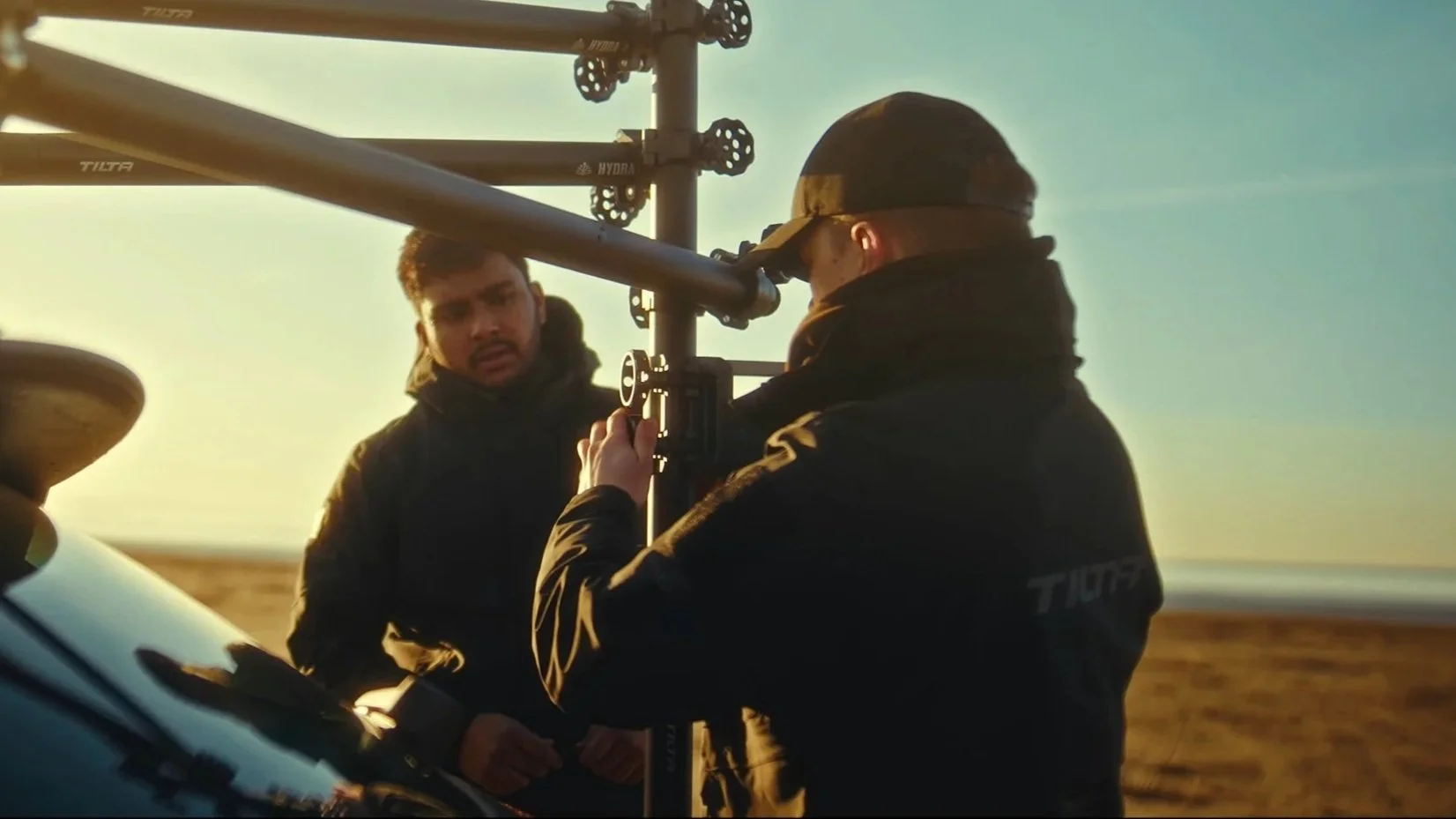 Two men in black jackets setting up filming equipment outdoors during sunset or sunrise, with a clear sky and open field in the background.