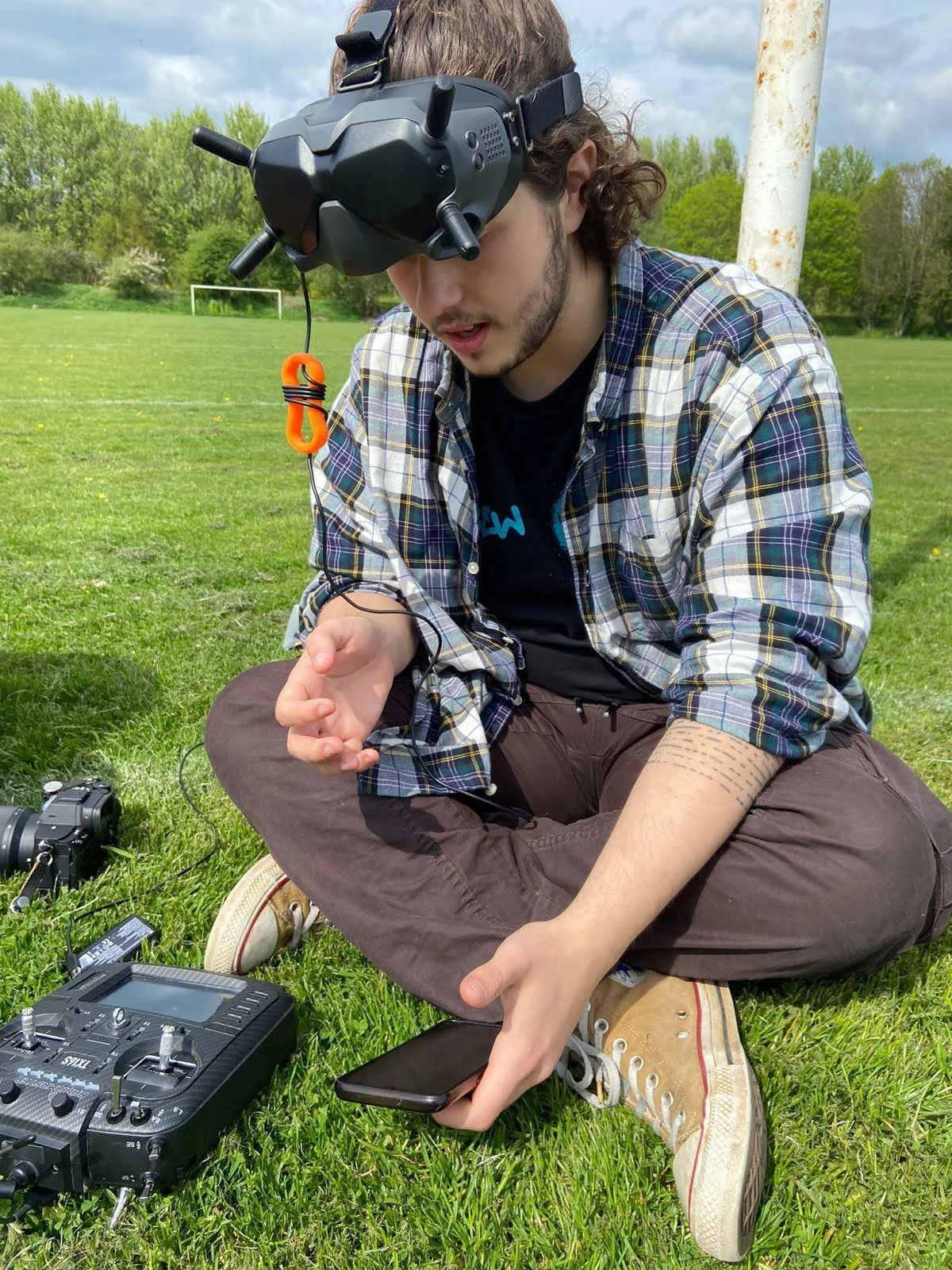 Stan Thornton, a young man sitting cross-legged on the grass with drone equipment, wearing FPV goggles, a plaid shirt, brown pants, and sneakers in an outdoor field.