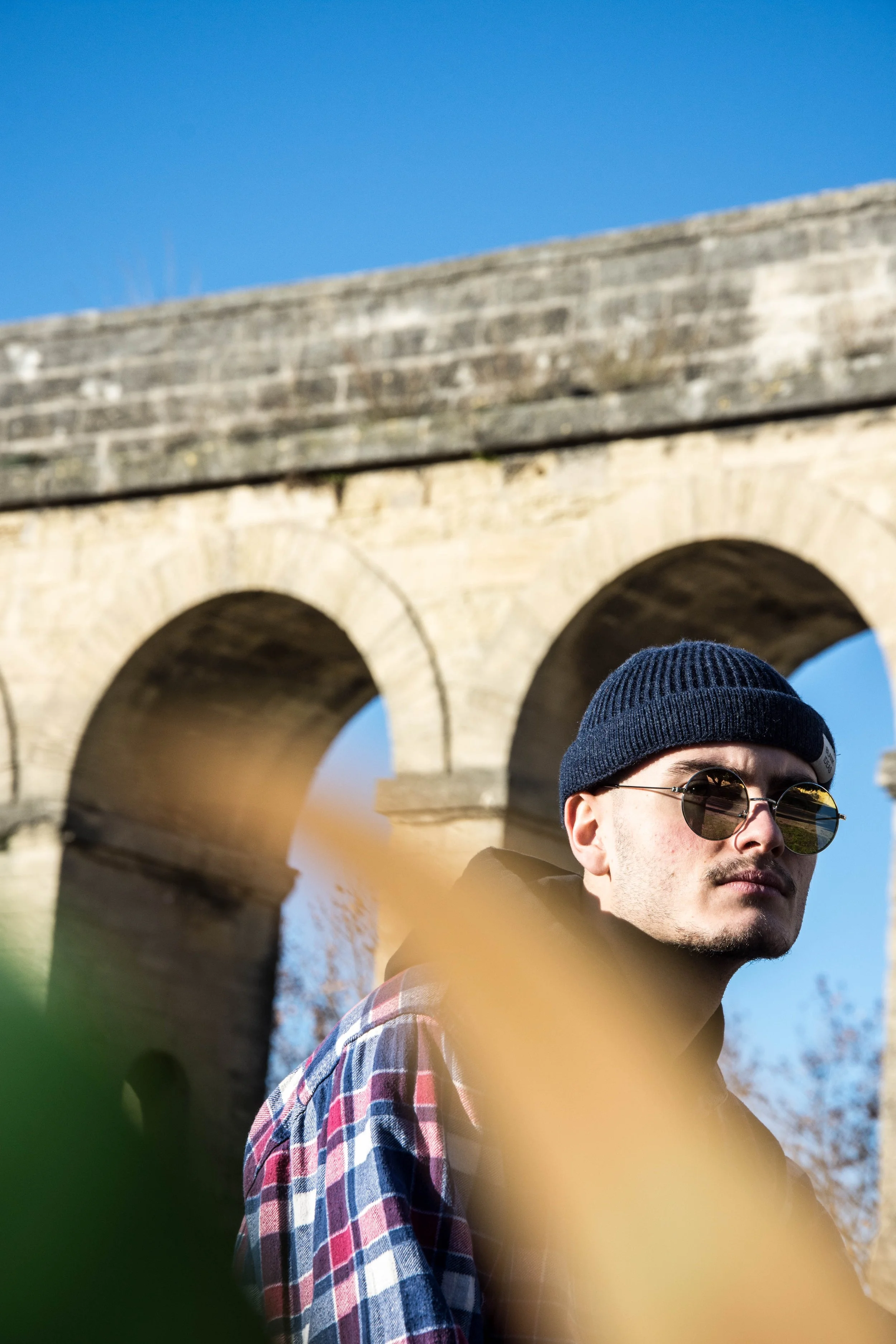 Grounded Media - Young man wearing sunglasses, a beanie, and a plaid shirt, standing outdoors near an old stone bridge against a clear blue sky - by stan thornton