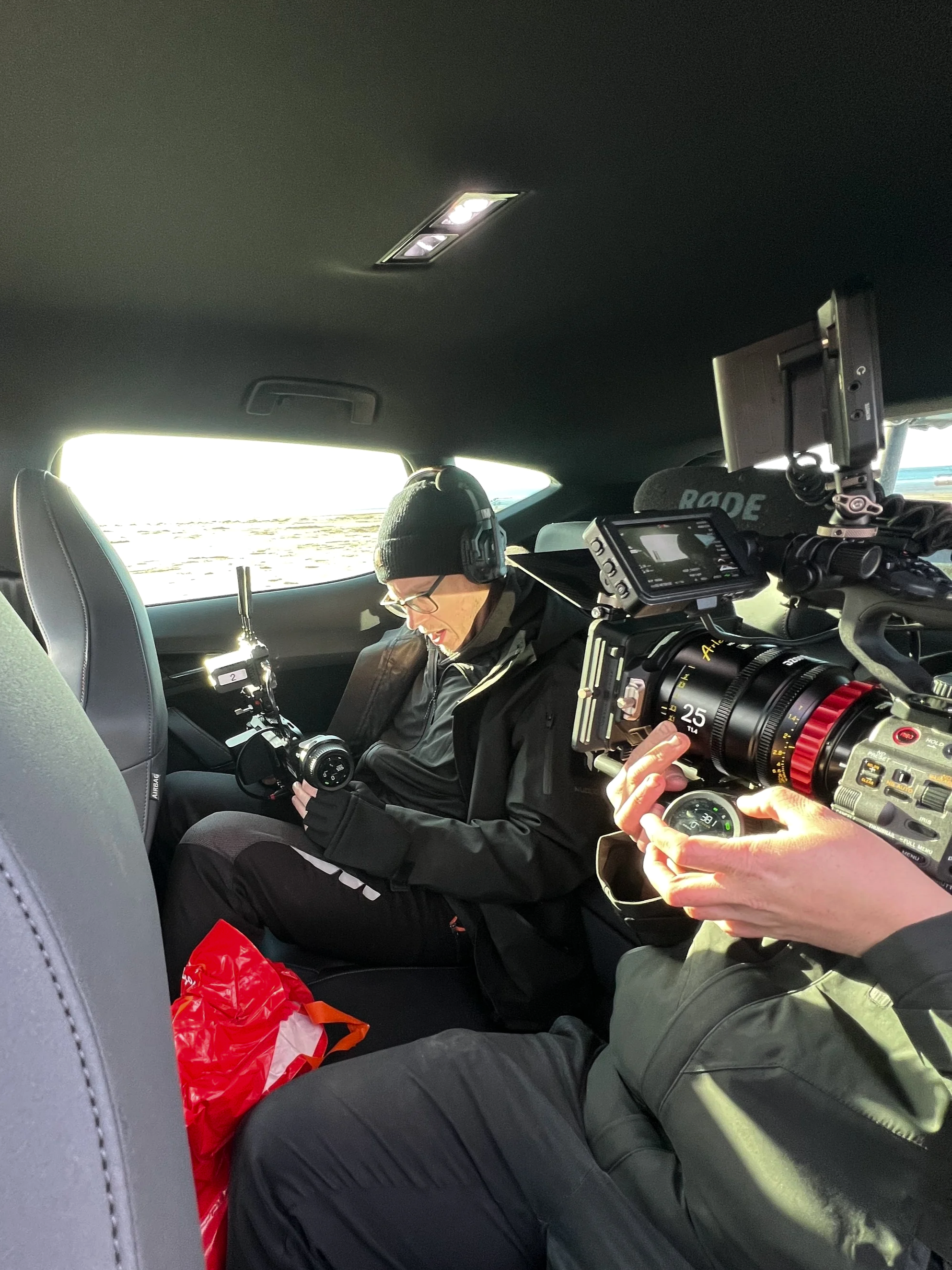 Two people inside a vehicle, one with a camera setup, filming another person sitting in the back seat, who is wearing headphones, glasses, a beanie, and casual clothing.
