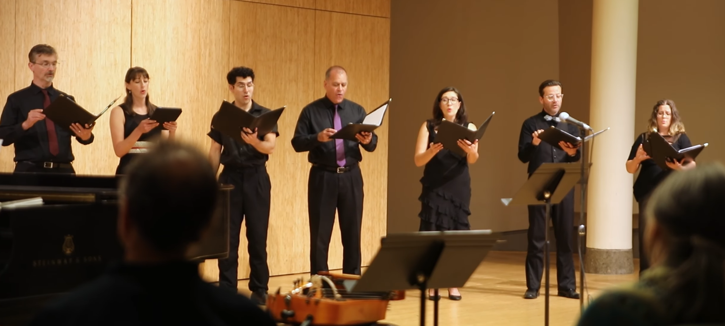 A group of seven singers standing on a wooden stage, holding open sheet music, performing in front of an audience. They are dressed in black clothing with some wearing purple accents, and there is a piano and a guitar visible in the scene.