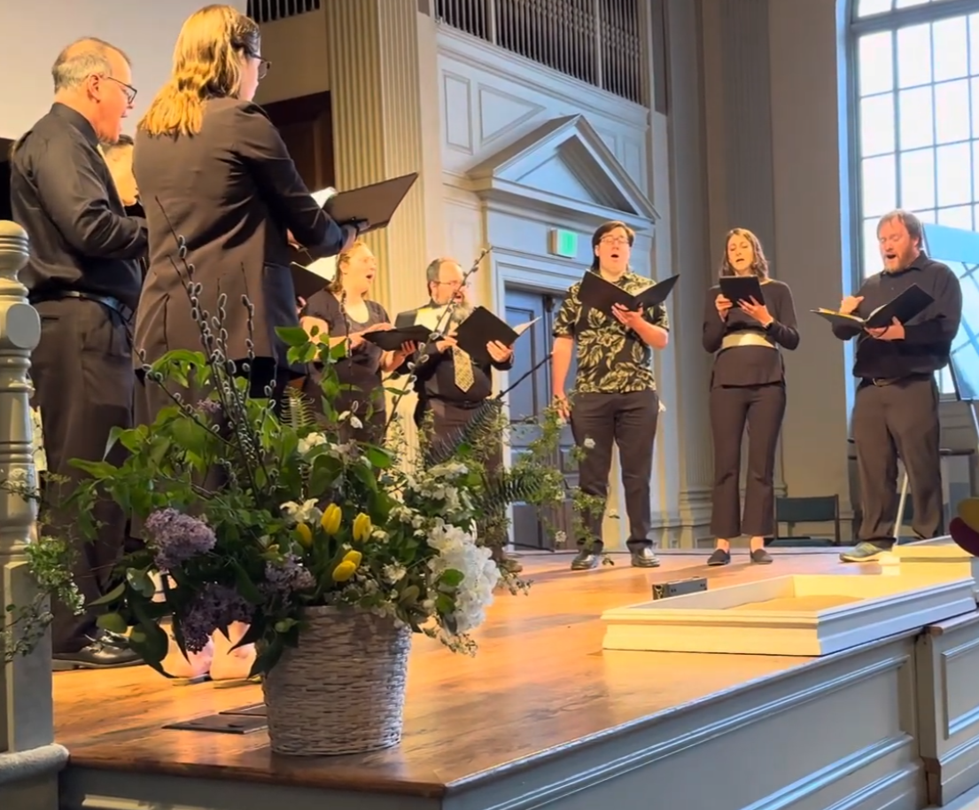 A choir of seven people singing on stage, holding sheet music, with a large flower arrangement in the foreground, in a church or chapel with tall windows and wood-paneled walls.