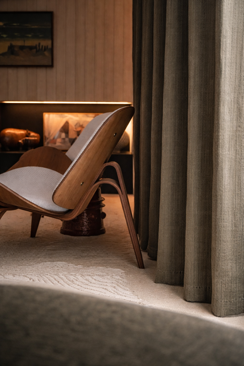 A cozy interior corner with a wooden lounge chair, curtains, a carpet, and warm ambient lighting.