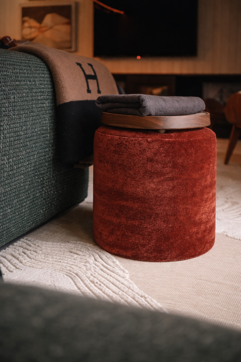 A cozy living room corner featuring a dark green textured sofa with a beige and black throw blanket, a red velvet ottoman, a wooden tray with a folded gray blanket on top, and a white textured rug on the floor.