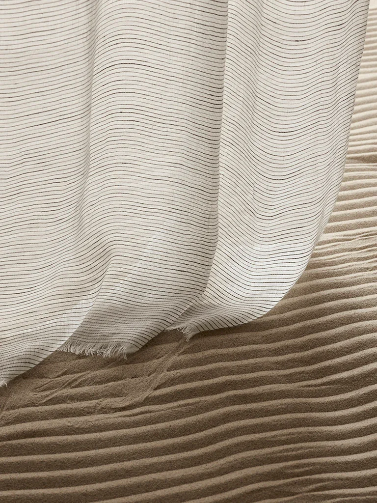 Close-up of a white curtain with thin black horizontal stripes, hanging over a textured, rippled sand surface.